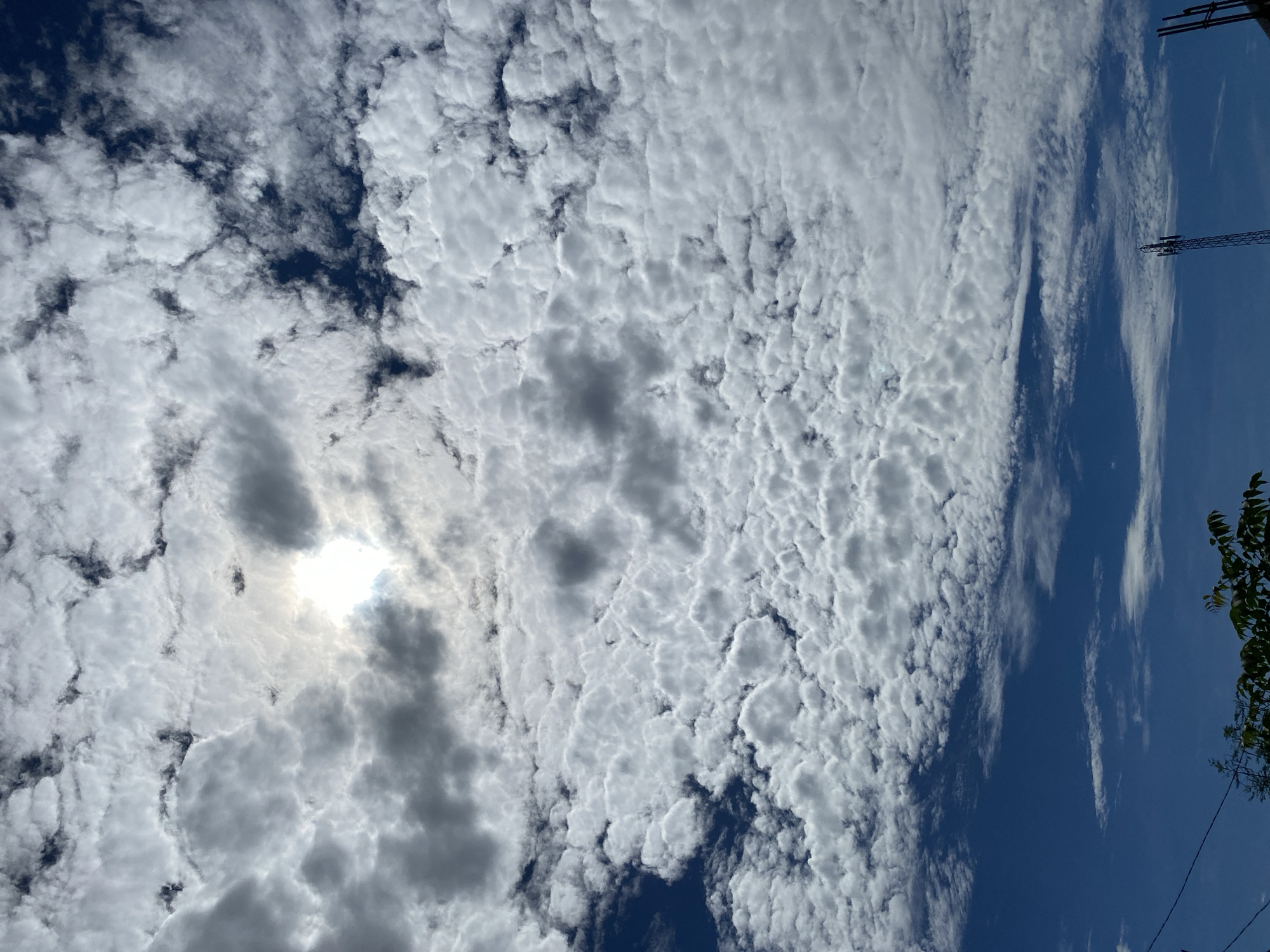 The image depicts a sky filled with a mix of cumulus and cirrocumulus clouds, with patches of dark blue sky visible. The sun is partially obscured by clouds, creating a bright, diffused light. The lower portion of the image shows a sliver of the horizon, with some trees and what appears to be a communication tower. This scene is likely from Banda Aceh, Indonesia, given the typical cloud formations and sky conditions.