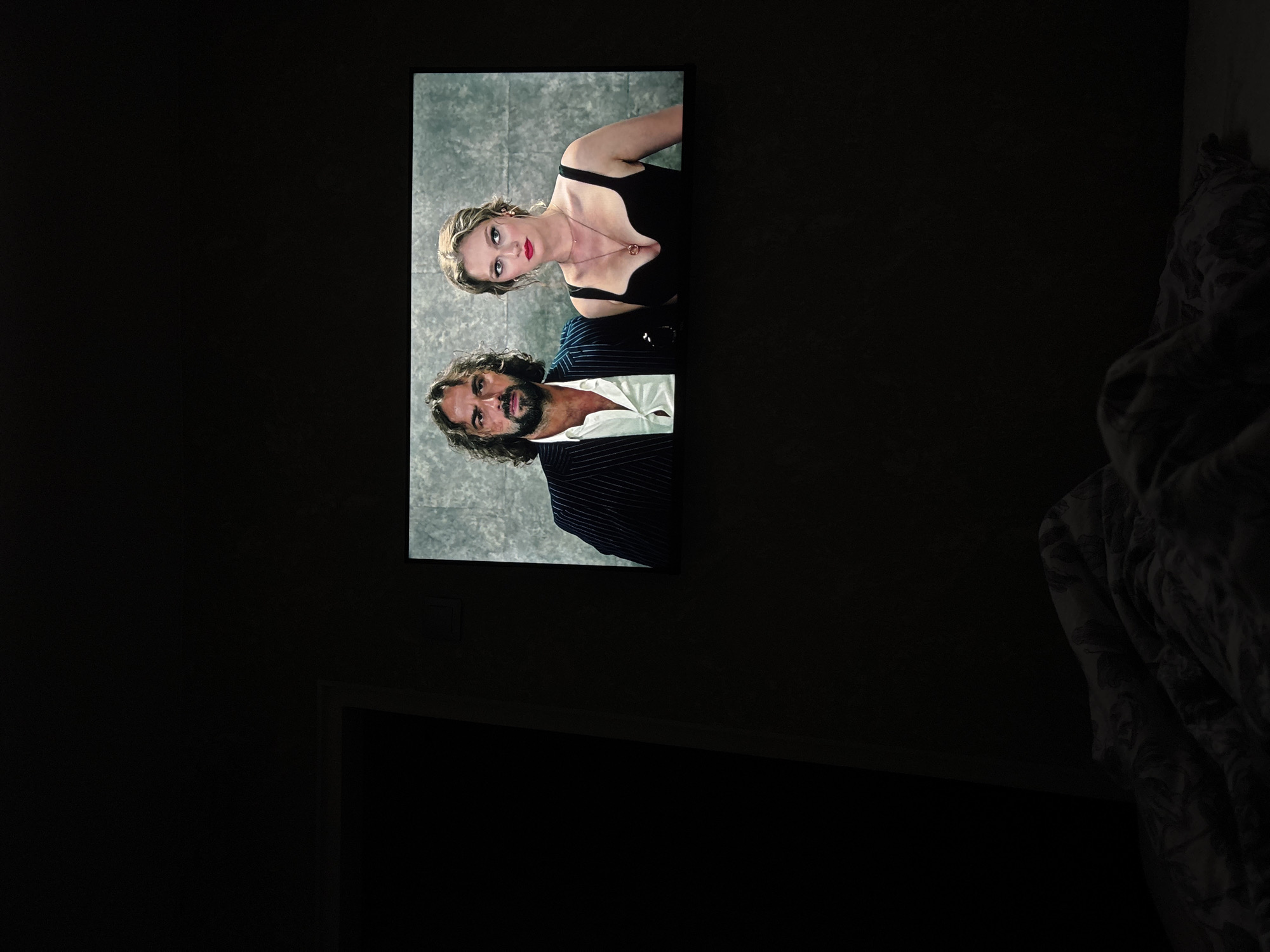 In a dimly lit room, a television screen displays a scene from a film featuring a man and a woman. The man, with long, dark, wavy hair and a beard, wears a pinstriped suit jacket over a white open-collared shirt. The woman, with blonde hair pulled back, wears a low-cut black dress and a necklace with a circular pendant. They are positioned side-by-side, looking towards the viewer. The background on the screen is a textured, light-colored wall. To the left of the television, a dark vertical shadow suggests a doorway or large object. In the foreground, a rumpled bed with patterned bedding is visible, shrouded in darkness. The overall scene implies the viewer is in a bedroom, watching television in low light conditions.