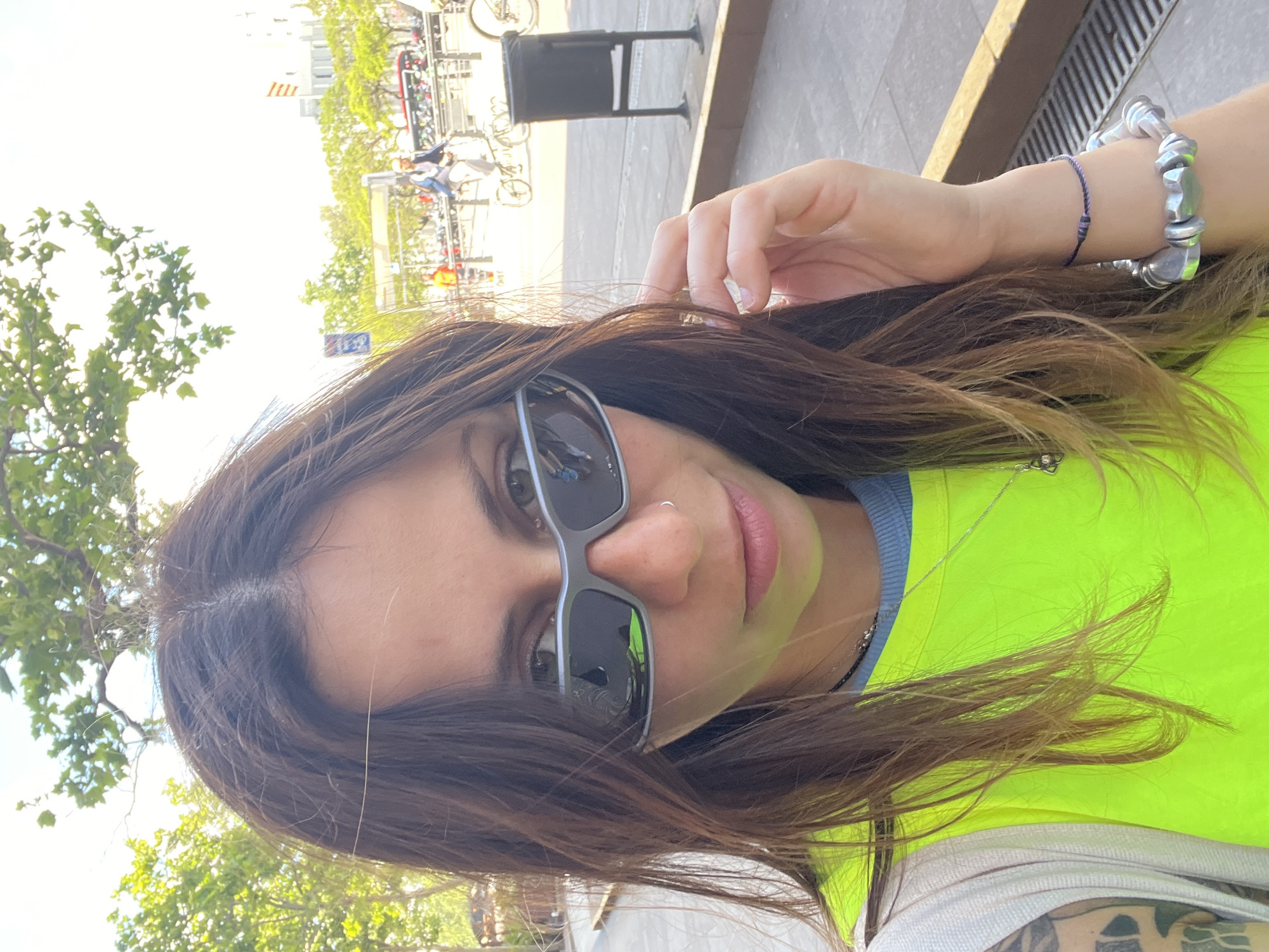 A selfie captures a woman with long brown hair, wearing sunglasses, and a bright neon yellow top. She also sports a silver bracelet and a blue string bracelet on her left wrist. The setting appears to be outdoors, possibly in a city. In the background, blurry images of bicycles, buildings, and greenery suggest an urban environment. Based on the architectural style and overall atmosphere, it is likely the location is in Barcelona, Spain.