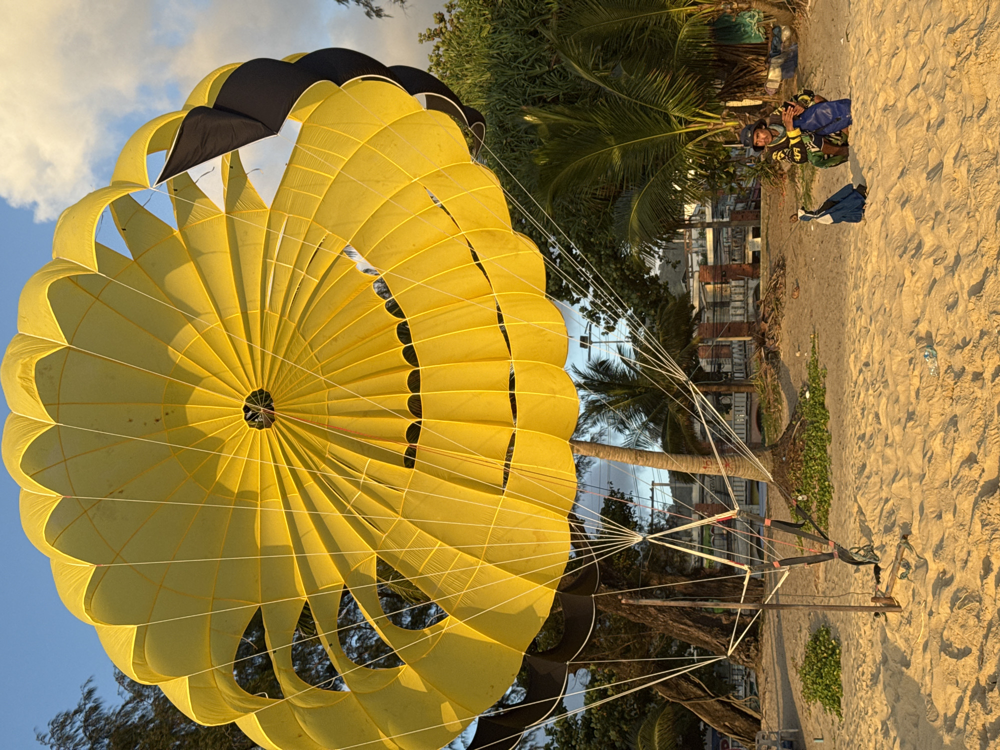 A yellow and black parasail is set up on a sandy beach in Karon, Thailand. The parasail is spread open and anchored to the ground with a wooden frame and harness. A man in a blue hat and a yellow and black patterned shirt is sitting on the sand next to a blue bag, holding a cell phone. Two other blue bags are next to him. Palm trees and other greenery are visible in the background, along with buildings and a cloudy sky. The sun is setting, casting a warm glow on the scene.