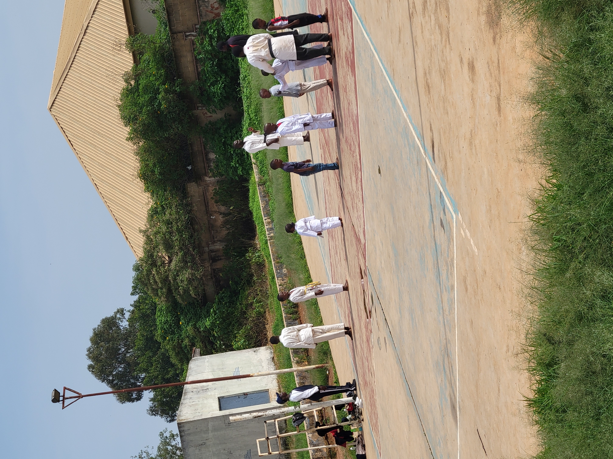 An outdoor, ground-level, slightly elevated, medium-wide shot captures a group of individuals, mostly children, in what appears to be a martial arts training session on a paved court. The court has distinct markings with blue and red lines. In the background, a building with a light brown, corrugated metal roof stands against a clear blue sky. Lush green trees and vegetation surround the area, with a grey, concrete building visible to the left, near a metal pole with a floodlight fixture.
The primary subjects are a group of individuals dressed in white karate uniforms, some with colored belts. Several adults, also in martial arts attire, appear to be instructors. One adult male in a white uniform with a black belt stands in the center of the group, seemingly instructing. Children are positioned in rows, facing the instructors or observing. A ladder-like structure with bags and shoes at its base is situated to the left. The ground shows some wear and tear with patches of darker concrete and water marks. The overall scene suggests an organized outdoor activity.