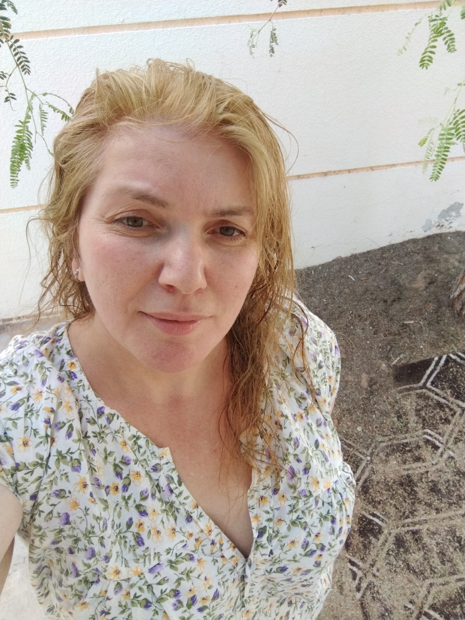 A middle-aged woman with blonde hair and fair skin is posing for a selfie outdoors. She is wearing a floral print dress. The background consists of a white wall with some greenery hanging from it. The ground appears to be covered in gravel or small stones. The location is possibly Al Mas'oudi, United Arab Emirates. A geometric pattern is visible on the ground, likely paving stones.