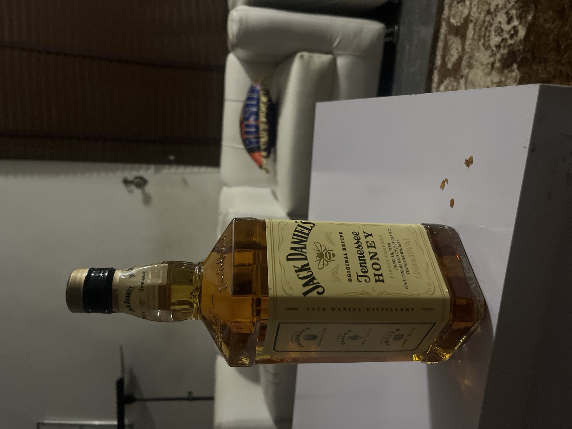 A bottle of Jack Daniel's Tennessee Honey liqueur is on a white table. The bottle is oriented horizontally. The table is in front of a white couch with a decorative pillow and a patterned rug. The location is not possible to discern from the image.