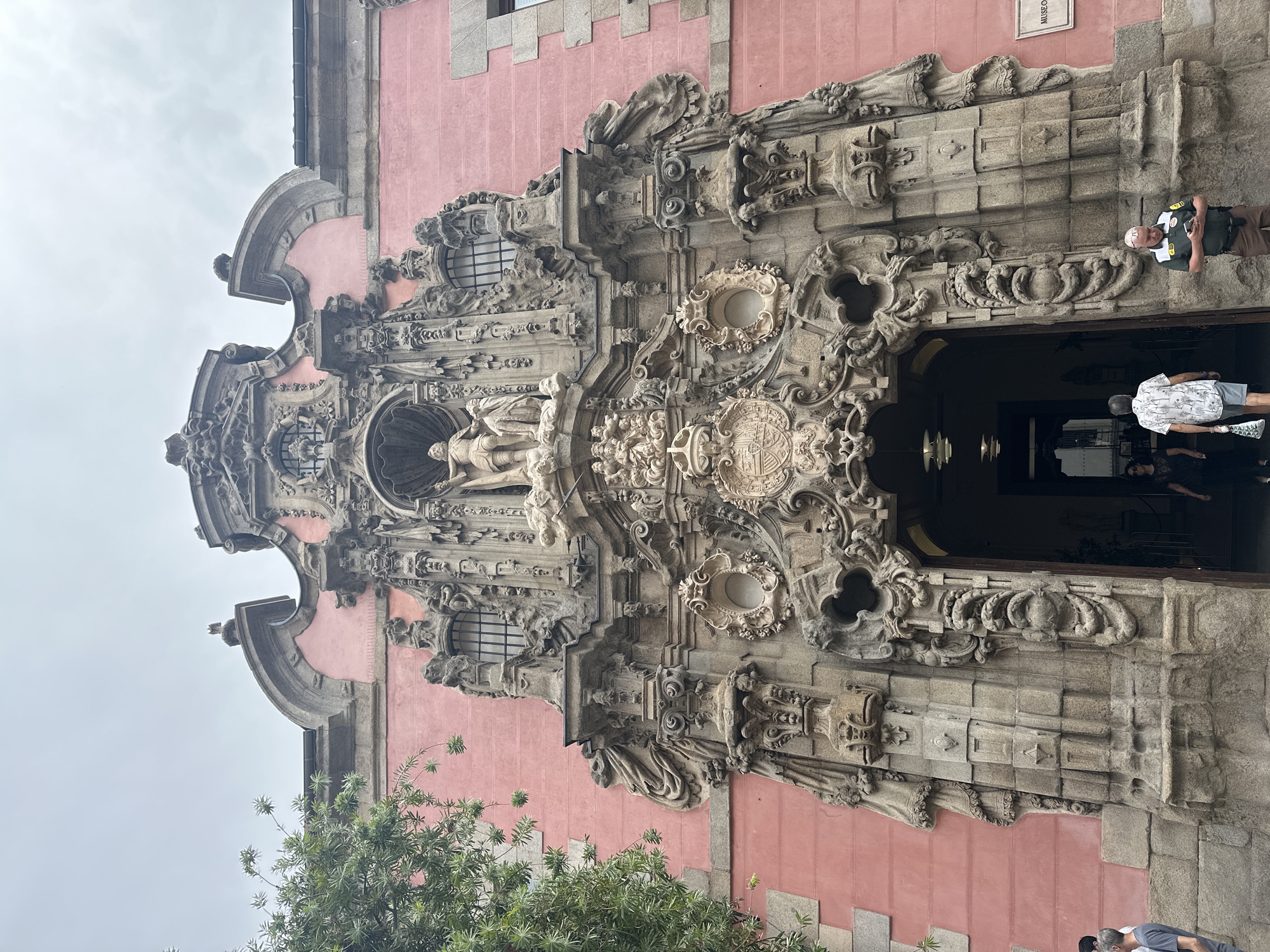 The image shows the ornate facade of a building in Madrid, Spain, specifically the entrance to the Museo Sorolla. The building's exterior is painted a vibrant pink, contrasting with the grey stone and intricate carvings that surround the central doorway. The entryway is framed by a highly decorated Baroque architectural element featuring statues, reliefs, and elaborate scrollwork. Above the entrance, a statue of a figure is prominently displayed. Several people are visible near the entrance, some entering the museum and others standing outside. The sky is overcast, suggesting a cloudy day. A lush green tree is partially visible on the left side of the frame.
