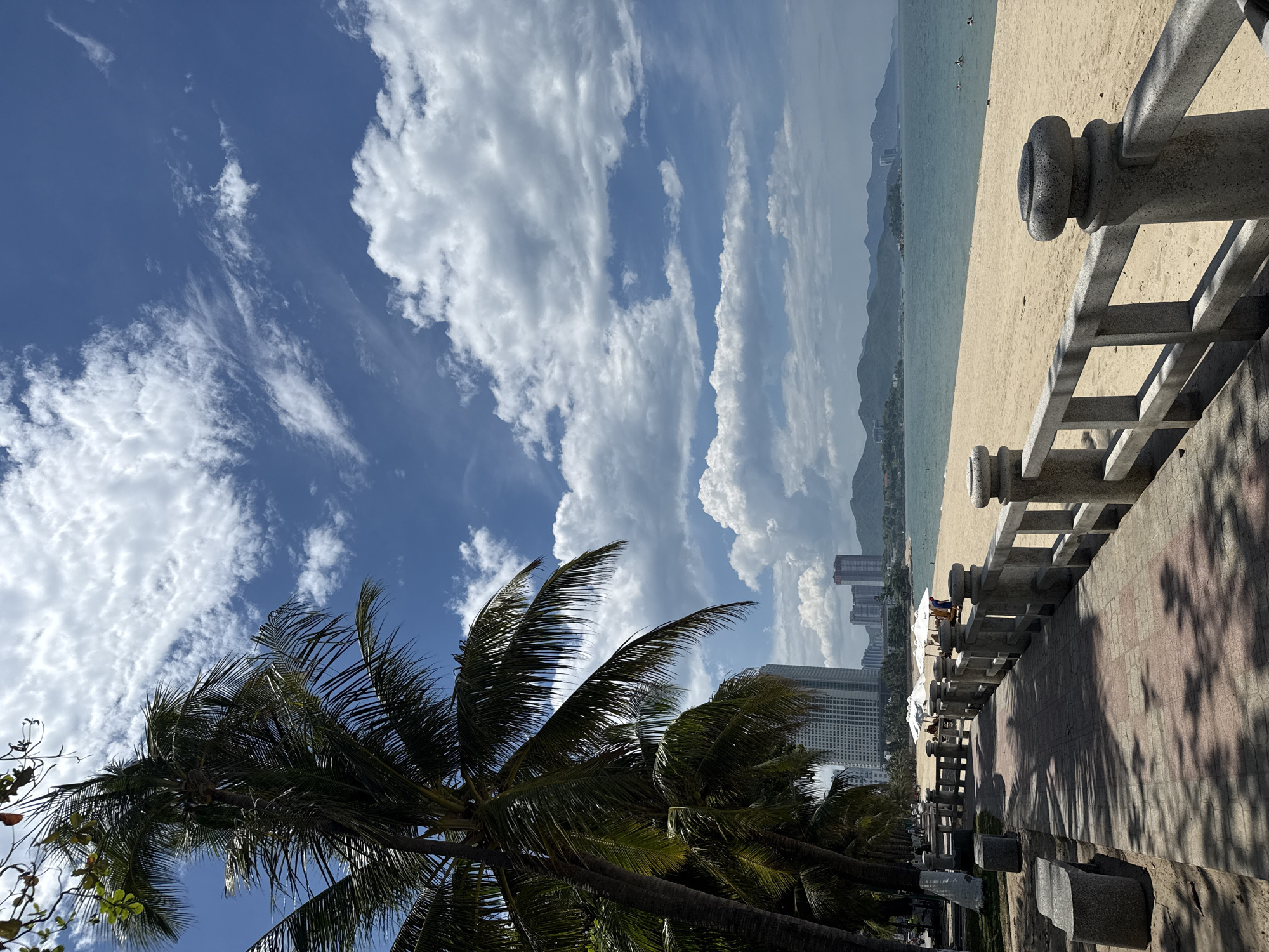 The image depicts a sunny day on a beach in Nha Trang, Vietnam. A paved walkway runs parallel to the sandy beach, bordered by a stone railing with decorative tops. Several palm trees with lush fronds are visible along the walkway, casting dappled shadows on the pavement. In the distance, high-rise buildings are clustered along the coastline, indicating an urban setting. The sea is a calm blue, with a few people swimming or wading in the water. The sky is a vibrant blue, adorned with fluffy white clouds. A mountainous landscape forms the backdrop on the horizon.