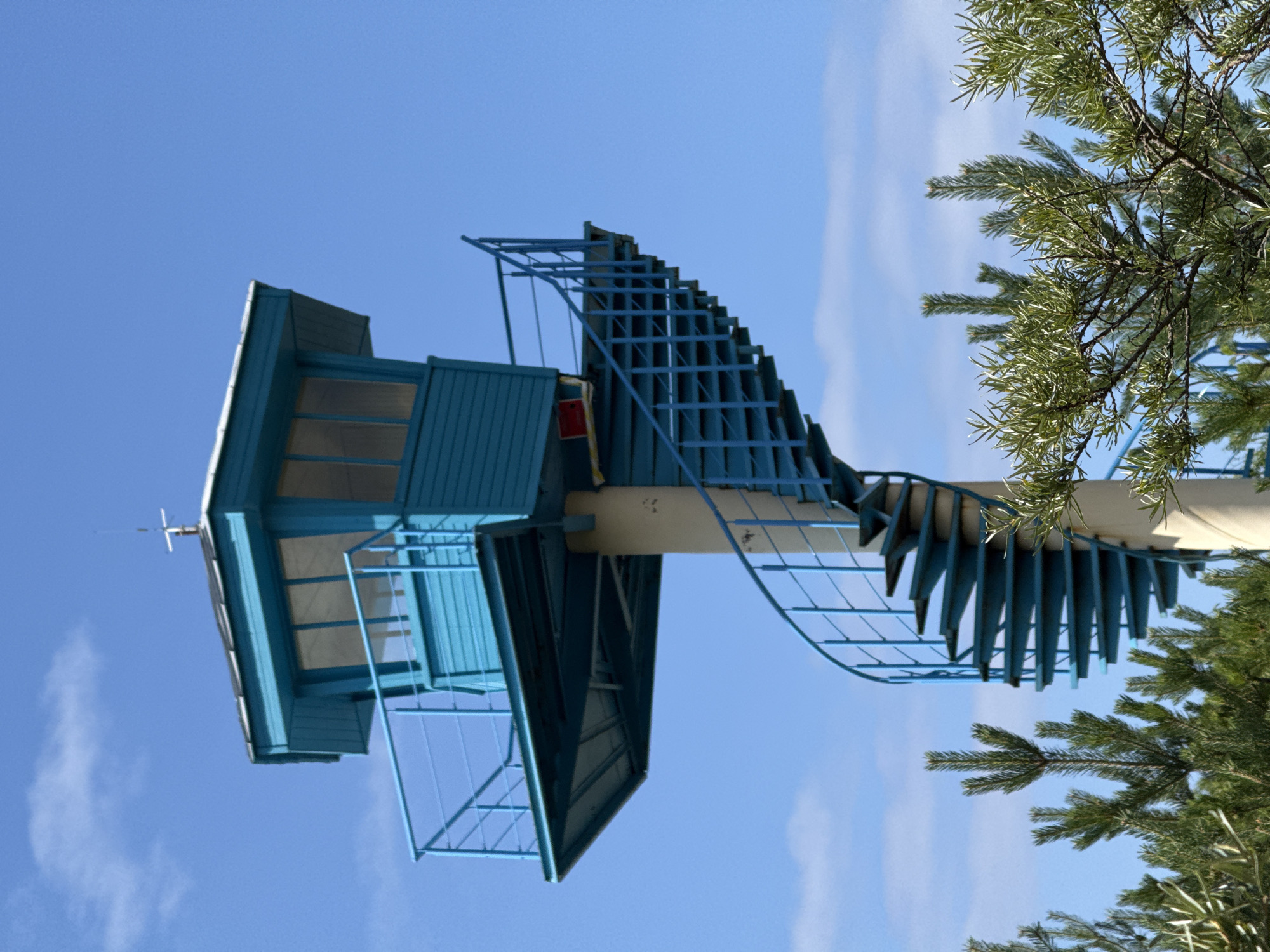This is a waist-up, slightly low-angle, eye-level shot of a bright blue lifeguard or observation tower with a spiral staircase leading up to it. The tower has a white roof and large windows. The staircase is also blue and wraps around a concrete pillar. In the foreground, there are out-of-focus green pine trees and some other leafy branches. The sky is clear blue with a few wispy white clouds. The observation tower is located on the beach in Ventspils, Latvia.