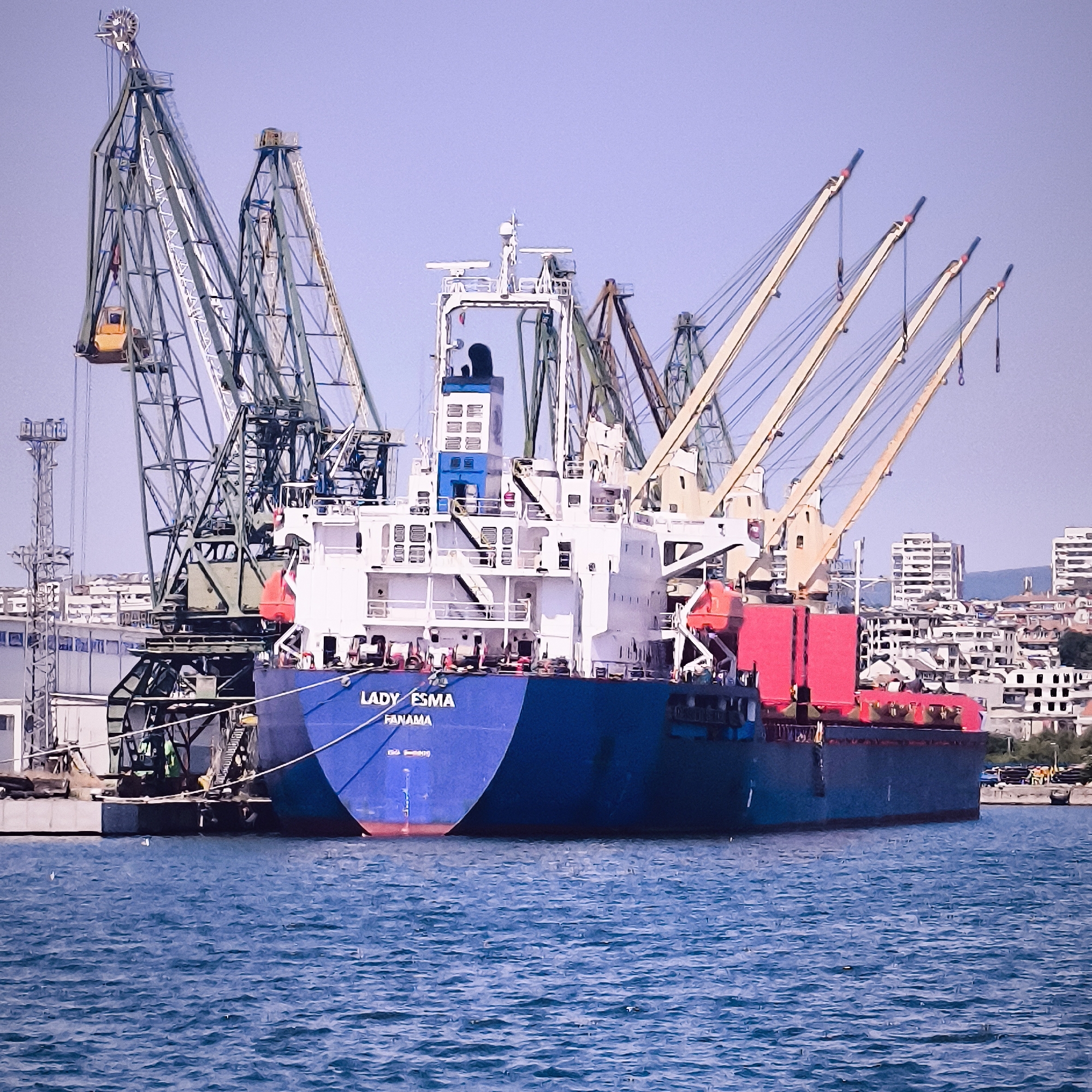 The image shows a large blue cargo ship named "LADY ESMA" with "PANAMA" written on its stern, docked at a port. The ship is positioned in front of several tall cranes, some of which are green and metal, while others have yellow booms. In the background, there is a city skyline with numerous buildings, some of which appear to be high-rise apartments. The scene is set against a clear, pale blue sky. The water in the foreground is a deep blue, with visible ripples. The ship appears to be in the process of being loaded or unloaded, as indicated by the presence of the cranes. The location is identified as Varna, Bulgaria, likely within the Port of Varna.