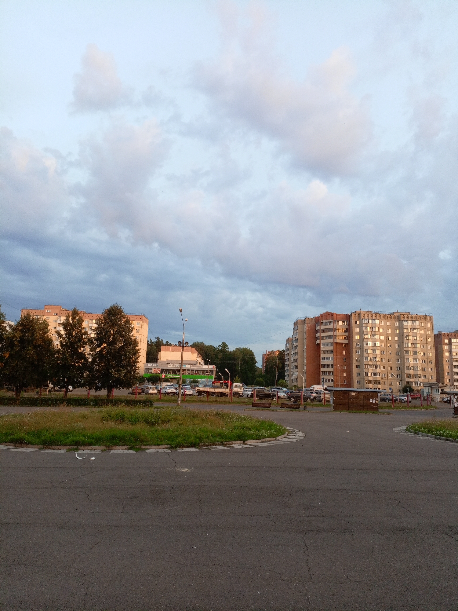 The image depicts a street scene in Zheleznogorsk, Russia. The foreground shows an asphalt road with visible cracks and a white line marking a curve. Beyond the road, there is a strip of green grass and vegetation. In the background, several multi-story apartment buildings are visible. On the left, three tall trees stand in front of a beige building with green and red accents. Further to the right, a larger, predominantly grey apartment building with orange brick details dominates the skyline. Several cars are parked along the road and in front of the buildings. The sky above is partly cloudy, with a mix of grey and white clouds against a pale blue backdrop. A street lamp with multiple light fixtures is present on the right side of the image. The scene appears to be captured during the daytime, possibly late afternoon or early evening, given the lighting.