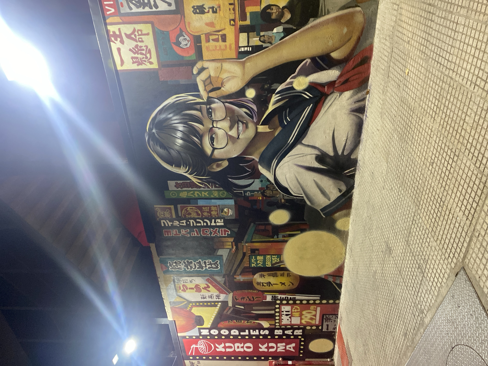 The image shows a mural on a wall, likely in an urban setting. The mural features a large portrait of a young woman with glasses and a Japanese school uniform. The background includes neon signs and other Japanese text, possibly indicating a restaurant or a cultural district. The mural also contains the words "KURO KUMA NOODLES BAR." A tiled sidewalk is visible adjacent to the mural. A bright light is seen in the left portion of the frame. I cannot confirm the location in the city of Buenos Aires, Argentina based on visual information alone.