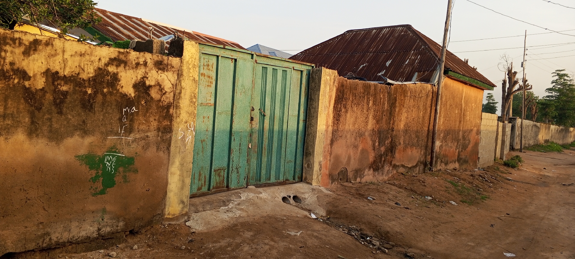 The image depicts a street scene in Jalingo, Nigeria, with a focus on a weathered, ochre-colored concrete wall and an adjacent green metal gate. The wall exhibits patches of dark discoloration and weathering, with some graffiti scribbled in white and green paint. Visible text on the wall includes "Ma a IF" and a green spray-painted area with the words "IMENENS". The green metal gate is of a ribbed design and appears to be slightly rusted. Two concrete drainage pipes are visible exiting the ground near the gate. Beyond the wall and gate, corrugated metal roofs of buildings are visible, along with a wooden utility pole and power lines. The ground is a mixture of dirt and sparse vegetation. The scene appears to be an outdoor environment, possibly in a residential or mixed-use area of the city.