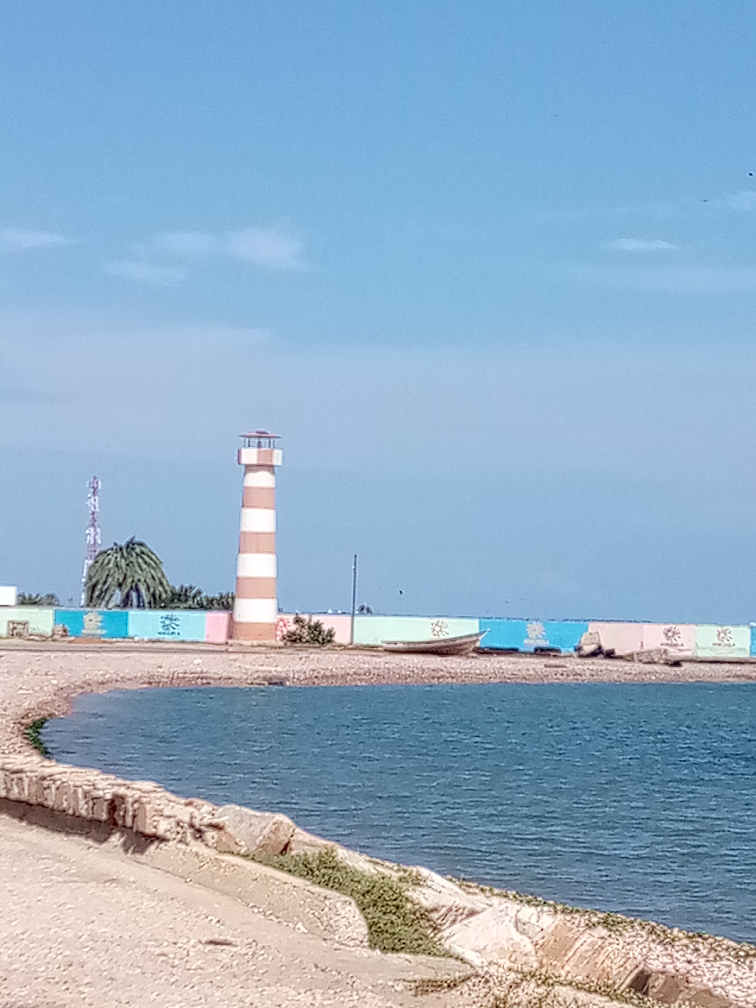 The image shows a lighthouse with alternating brown and white stripes, situated on a rocky shoreline. The lighthouse is tall and cylindrical, with a domed top. In the background, there is a colorful wall with various shades of blue, pink, and green. Several boats are pulled up onto the shore in front of the wall, and some vegetation is visible along the shoreline. A cell tower and a palm tree are also present in the distance. The sky is a clear blue with a few scattered clouds. The water in the foreground is a deep blue. The scene appears to be a coastal area in the city of Punta de Piedras, Venezuela, likely a port or a public beach area.