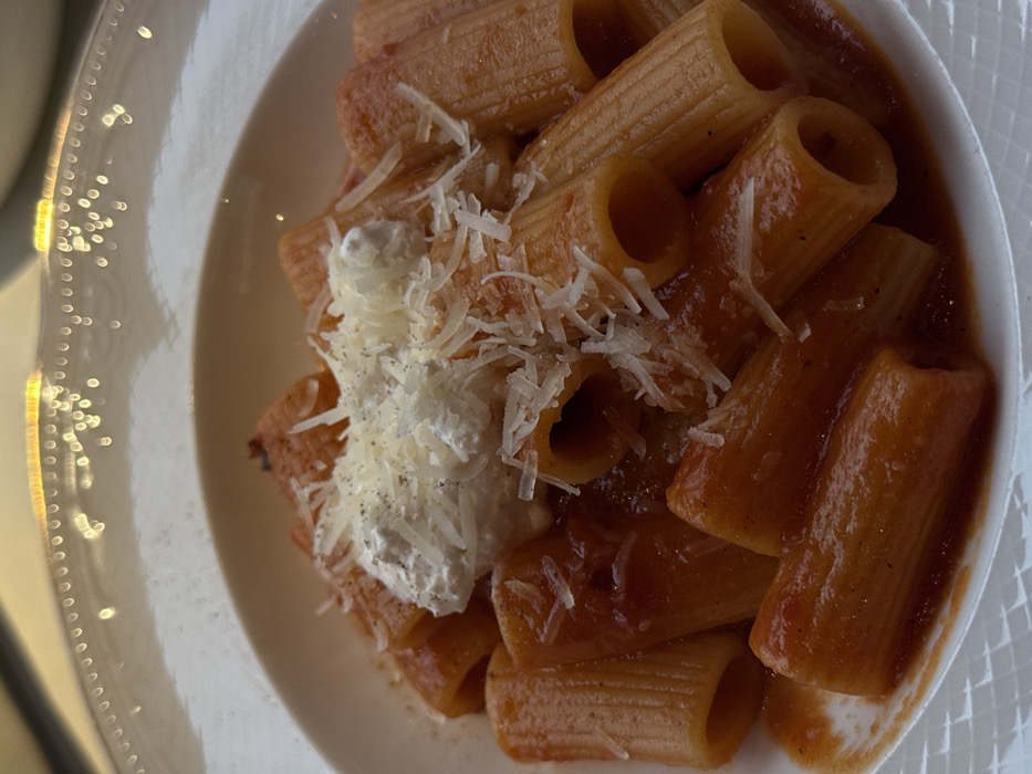The image displays a close-up shot of a serving of pasta in a white, shallow bowl or deep plate. The pasta appears to be rigatoni or a similar large, tubular, ridged pasta, coated in a rich, reddish-brown tomato-based sauce. Many individual pieces of pasta are visible, some standing upright, showcasing their hollow centers.
Generously sprinkled over a portion of the pasta is a layer of finely grated or shredded white cheese, likely Parmesan or Pecorino Romano. Below this shredded cheese, there is a distinct dollop of a creamy white, softer cheese, possibly ricotta, which adds a contrasting texture and color to the dish. Small, dark specks, likely freshly ground black pepper, are visible on top of both the shredded cheese and the creamy white dollop.
The white ceramic plate has a decorative raised pattern, possibly a series of small, reflective dots or beads, along its rim, particularly visible on the left side where some bright, out-of-focus light reflections create a bokeh effect. The overall lighting suggests an indoor setting, possibly under artificial light, creating a warm and inviting presentation of the meal. No people or text are visible in the image. The focus is entirely on the food, highlighting its textures and toppings, indicating a prepared meal ready for consumption.