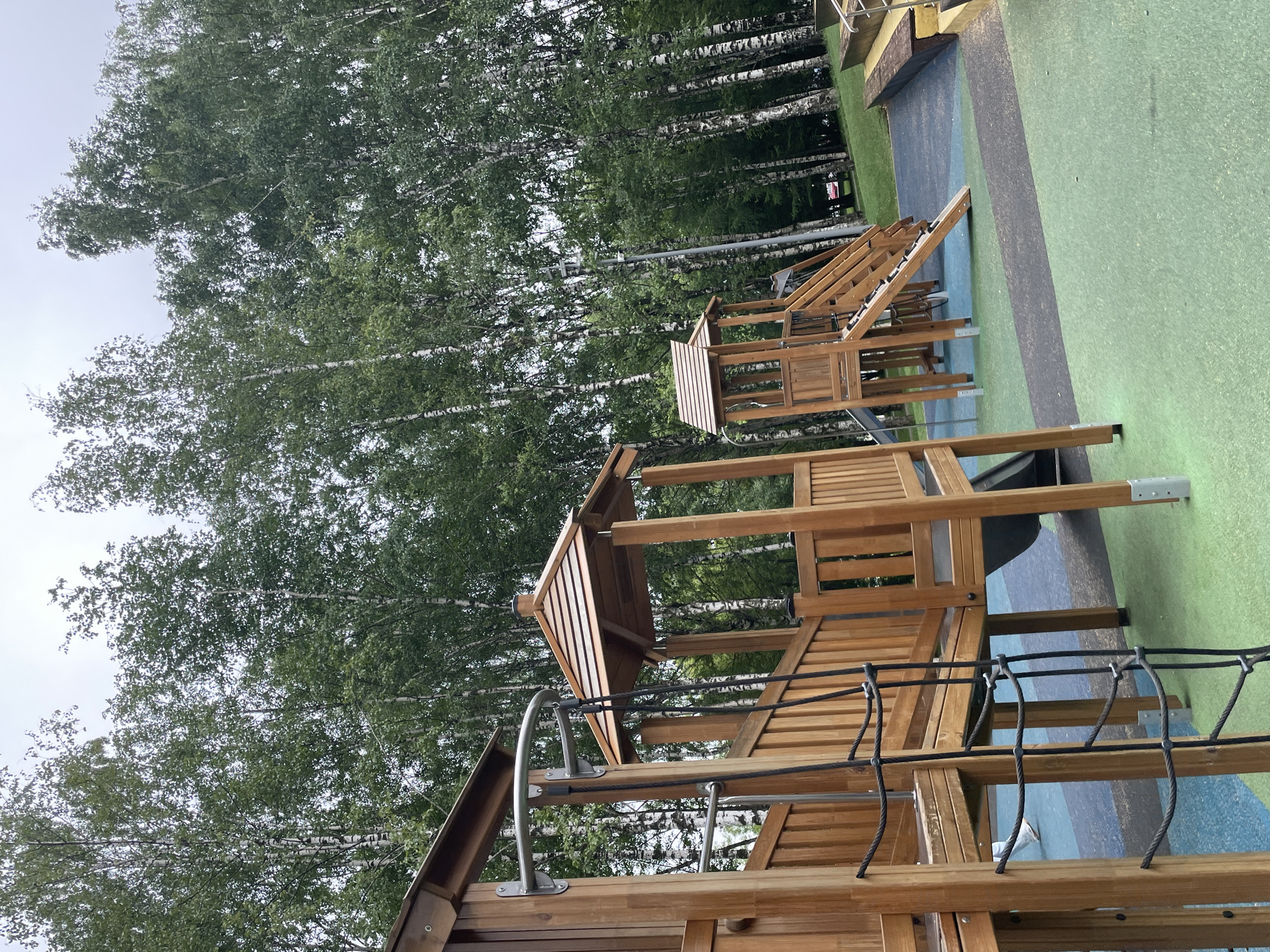In Kirov, Russia, a wooden playground structure stands in a park. The playground features multiple levels, a rope ladder, slides, and a climbing ramp. The ground is covered with a multi-colored rubberized surface in shades of green, blue, and black. Behind the playground, a dense forest of birch trees with white bark and green foliage creates a natural backdrop. The sky is overcast. No people are visible interacting with the playground equipment.