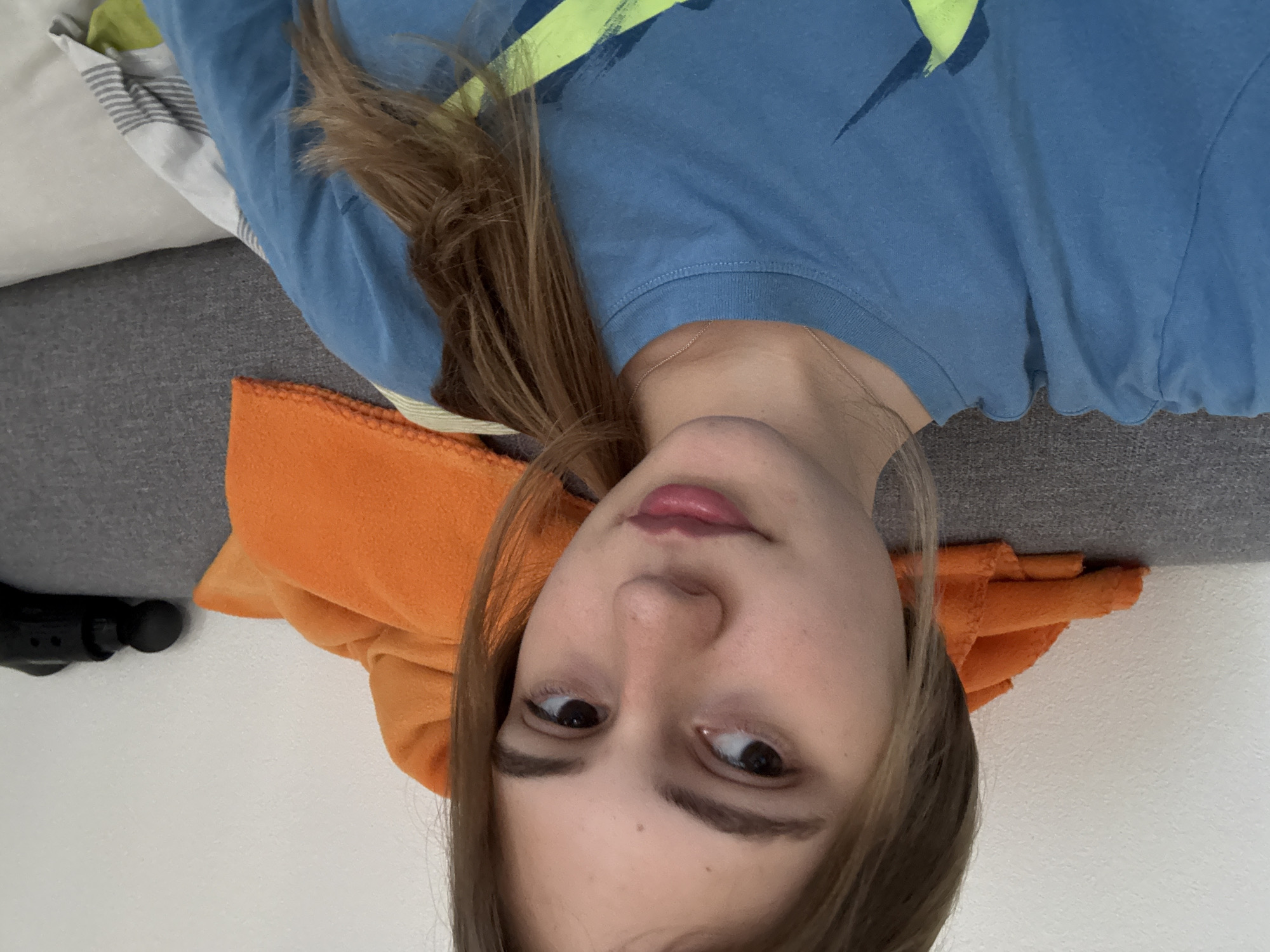 A young Caucasian woman with light brown hair, pulled back in a ponytail, is captured in a close-up, selfie-style shot. She is wearing a blue t-shirt with a graphic design. Her gaze is directed slightly off-camera to her left. In the background, an orange blanket is draped over a gray sofa, and a black electronic device is visible on the right side of the frame, resting on the sofa. The wall behind the sofa is a textured white. This image was taken indoors, in what appears to be a residential setting.