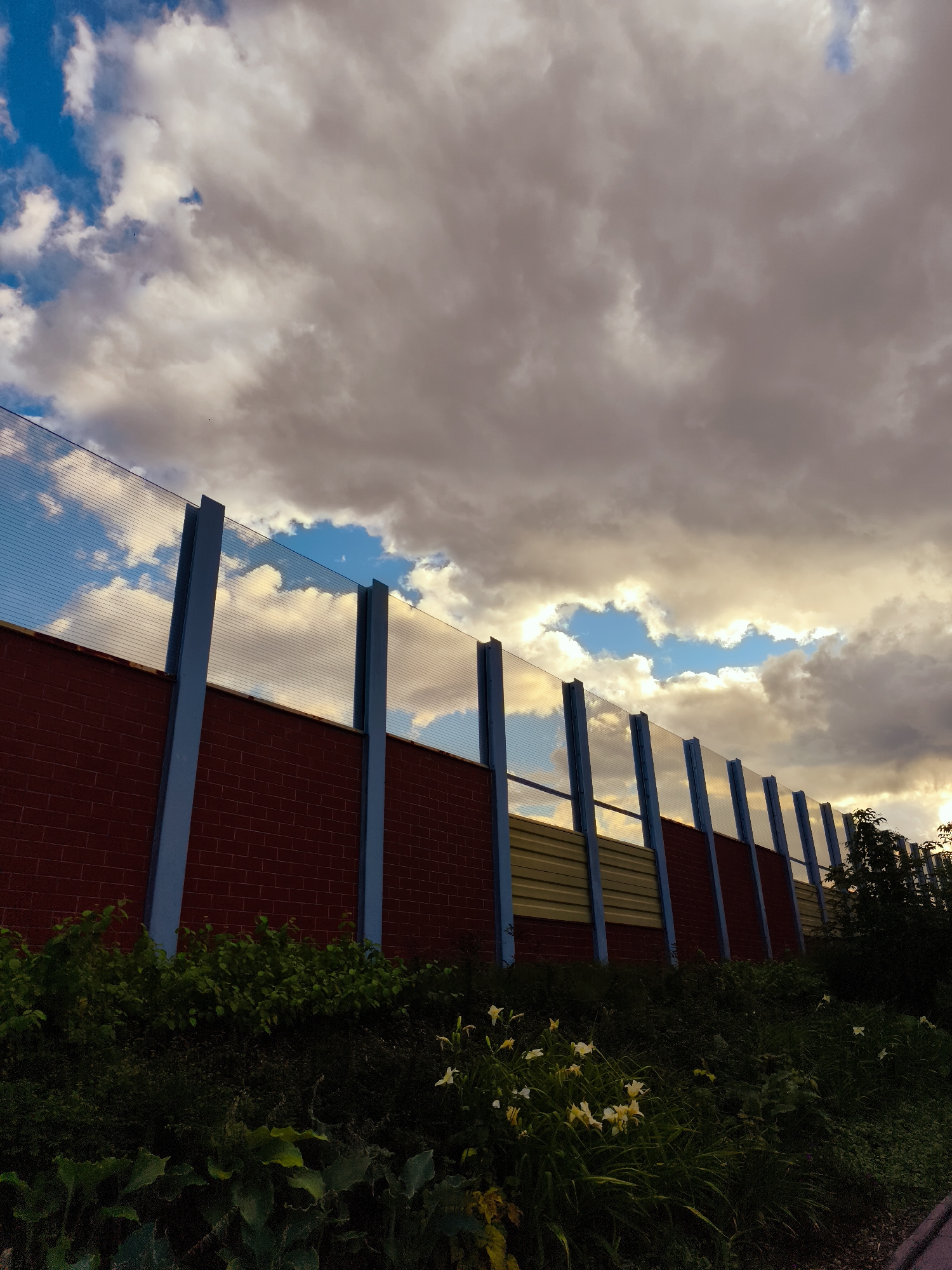 The image depicts a section of a sound barrier along what appears to be a road or industrial area, located in Gdańsk, Poland. The barrier is constructed with a lower section of red brick and an upper section of translucent corrugated material supported by blue metal posts. Beyond the barrier, a dense area of green foliage and some white flowers are visible in the foreground, suggesting a roadside or parkland setting. The sky is filled with dramatic clouds, illuminated by the warm glow of sunlight breaking through, indicating either sunrise or sunset. The overall scene is one of urban infrastructure contrasted with natural elements and a striking sky.