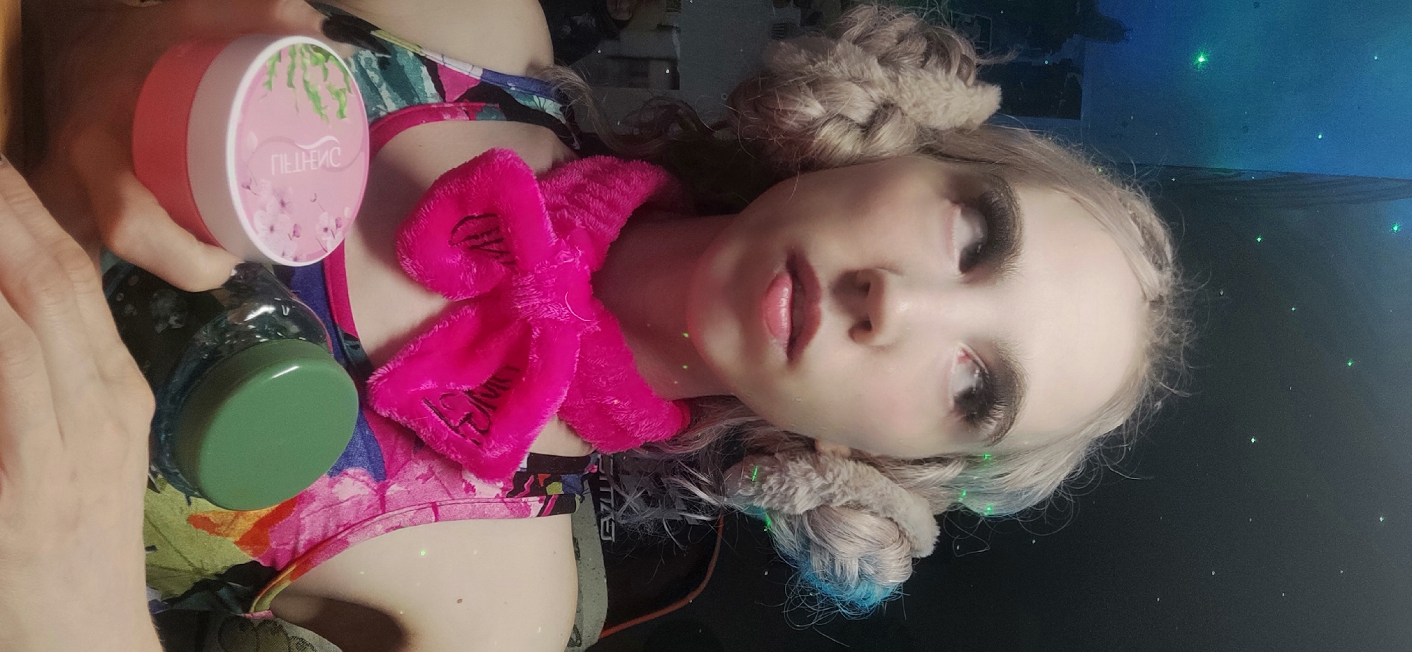 A person is holding two jars in a low-angle, selfie-style composition. The jar closest to the viewer is green with a dark liquid inside. Another jar is pink and white and has text on it that appears to be written in Russian. The person has light skin, light-colored hair that is braided, and a pink fuzzy bow around their neck. The person is wearing a multi-colored top. The background is dark, and there are green light beams in the background. The location and city are not determinable from the image.