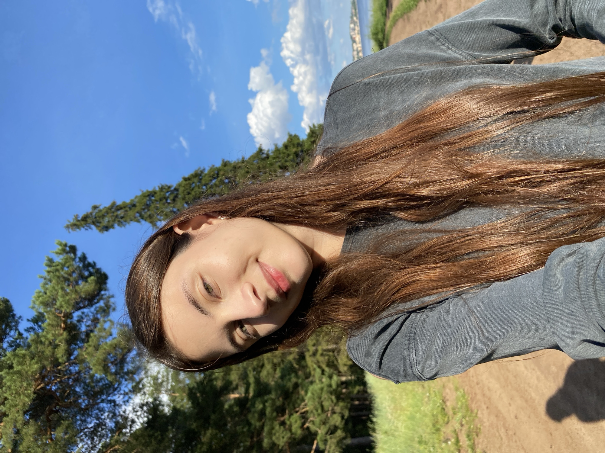 In Votkinsk, Russia, on a sunny day with a bright blue sky and scattered white clouds, a young woman with long, dark brown hair is taking a selfie outdoors. She is wearing a gray long-sleeved shirt. She is standing on a dirt path, with lush green grass and tall pine trees to her left. The background shows a body of water and a distant town or settlement. The light suggests it is late afternoon, casting a shadow of the woman on the path to her left.