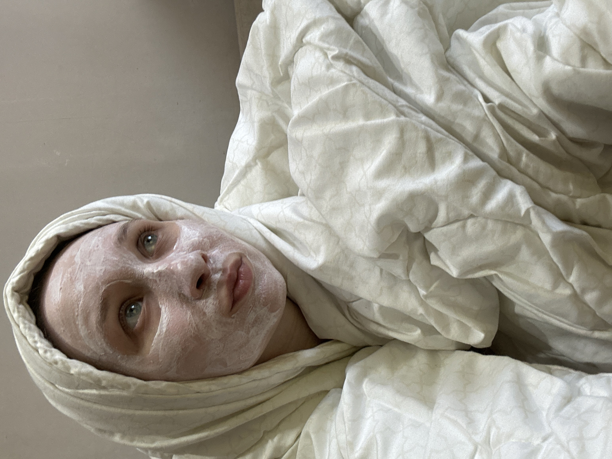 The image, taken in Chaikovsky, Russia, shows a young woman lying down and wrapped comfortably in a voluminous, light-colored comforter. Her face is covered with a thick layer of a white facial mask, contrasting with her visible light blue-green eyes, which are open and looking upwards. Her lips are slightly parted. The comforter, likely a duvet or blanket, is a soft off-white color with a subtle patterned texture, and is draped around her, including her head, giving a cozy and intimate feel. The background is a plain, light beige wall. The lighting is soft and natural, suggesting a relaxed daytime atmosphere. No text is visible. The scene evokes a sense of self-care and quiet relaxation.