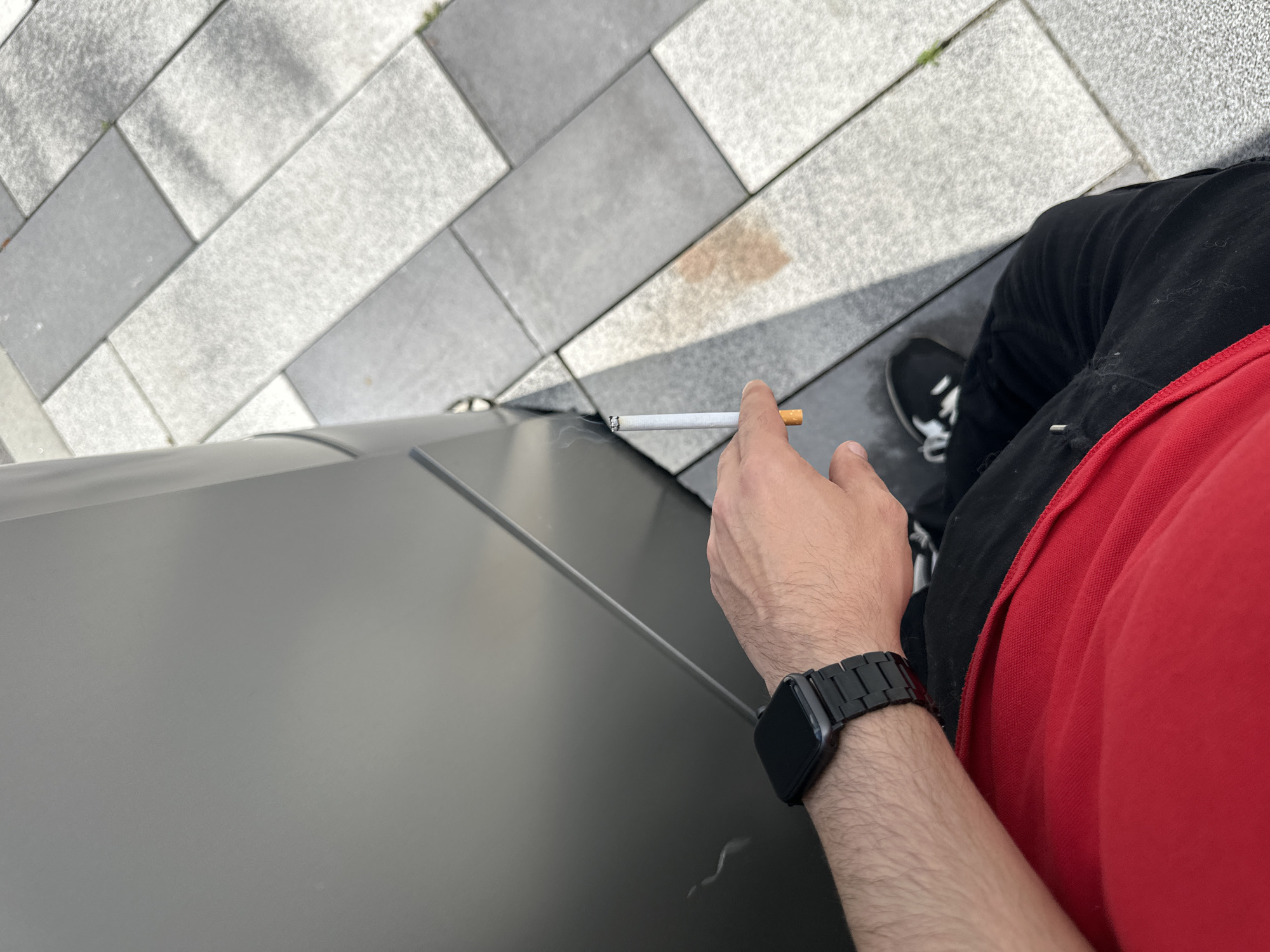 A high-angle, point-of-view shot shows a person holding a lit cigarette. The person is wearing a red shirt with black pants and black shoes that have white laces. On the person's left wrist is a black smartwatch. The person appears to be standing near a black, reflective surface, with gray and white concrete pavers in the background. There is no evidence of the location being Lovech, Bulgaria.