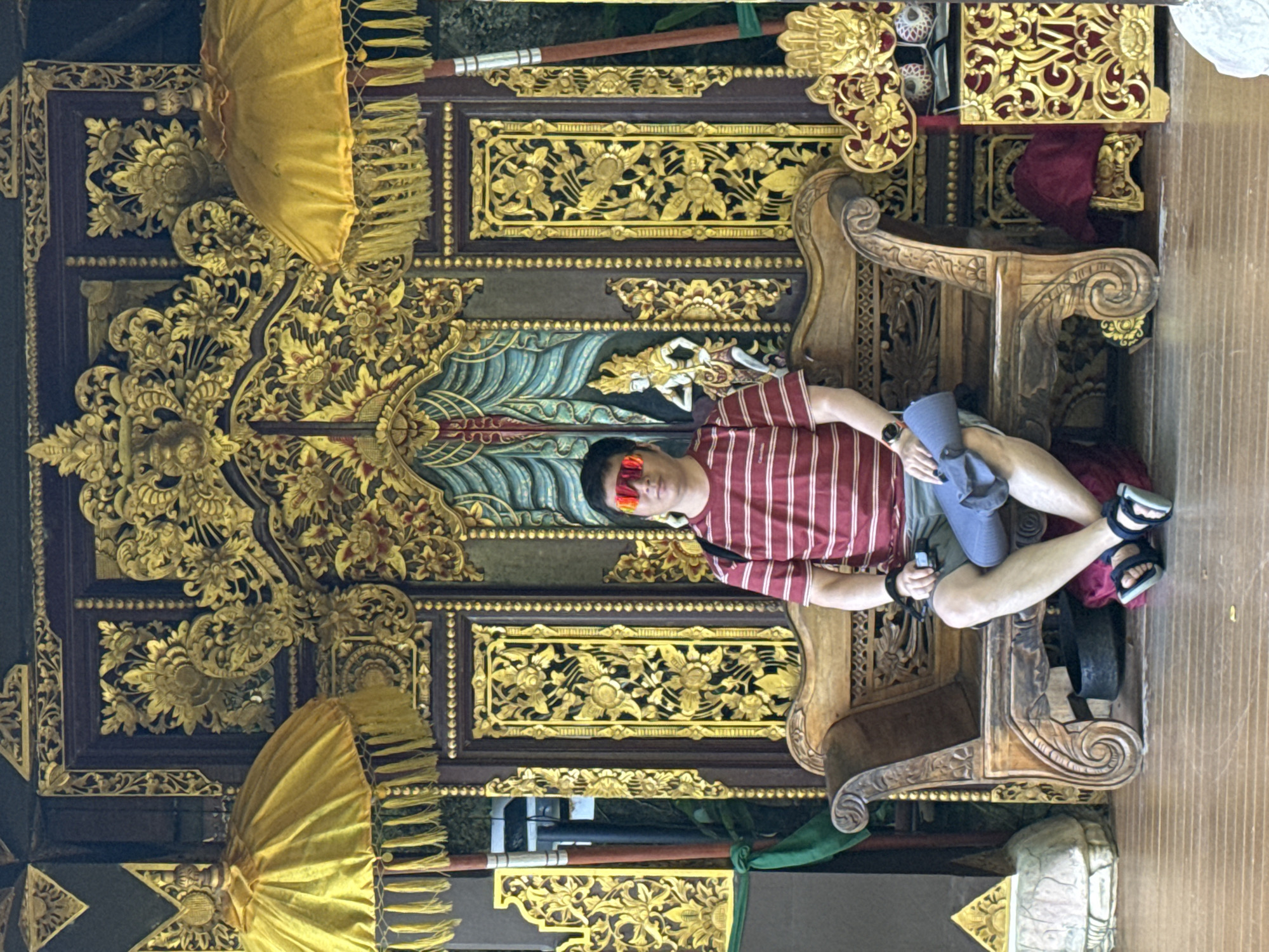 A full-length medium shot captures a person sitting on an ornate chair against an intricately carved backdrop. The individual, wearing a red and white striped shirt, light-colored shorts, red sunglasses, and black sandals, holds a rolled-up gray garment. They are seated on a wooden chair embellished with carvings. The backdrop features elaborate gold and black carvings, enhanced by two yellow umbrellas positioned at the top and bottom. The location is identified as Kuta Selatan, Indonesia.