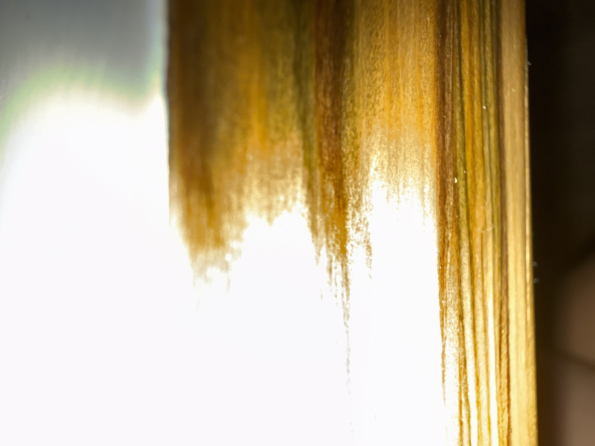 The image shows a close-up, abstract view of a golden, metallic surface. The surface appears to be textured with horizontal lines and is highly reflective, with bright white light dominating the upper left portion and transitioning into golden, brown, and greenish hues. There is a suggestion of a horizon line in the middle ground, where the reflective surface meets a darker, less distinct area. No people, specific objects, or discernible actions are visible. The location in Okhtyrka, Ukraine, cannot be determined from the provided image.