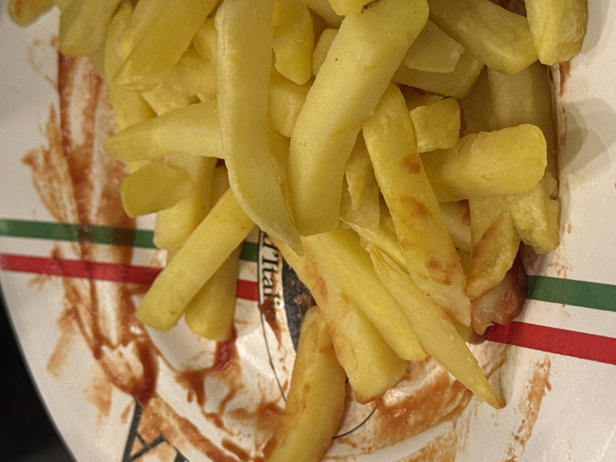 A close-up view depicts a pile of golden-yellow french fries served on a white, likely disposable, plate. The fries are thick-cut and appear lightly cooked, with some exhibiting a slightly crispier texture and deeper golden color. Numerous red ketchup smears and residue are visible on the white surface of the plate, indicating partial consumption. The plate features a decorative design with vertical stripes in red, white, and green, reminiscent of the Italian flag. A partial circular logo is discernible on the plate, revealing the text "r Ital" against a dark background, strongly suggesting branding such as "Pizzeria Italiana." No people are visible. The background is dark and out of focus. The scene is situated within a casual Italian-themed fast-food establishment or pizzeria in Sofia, Bulgaria.