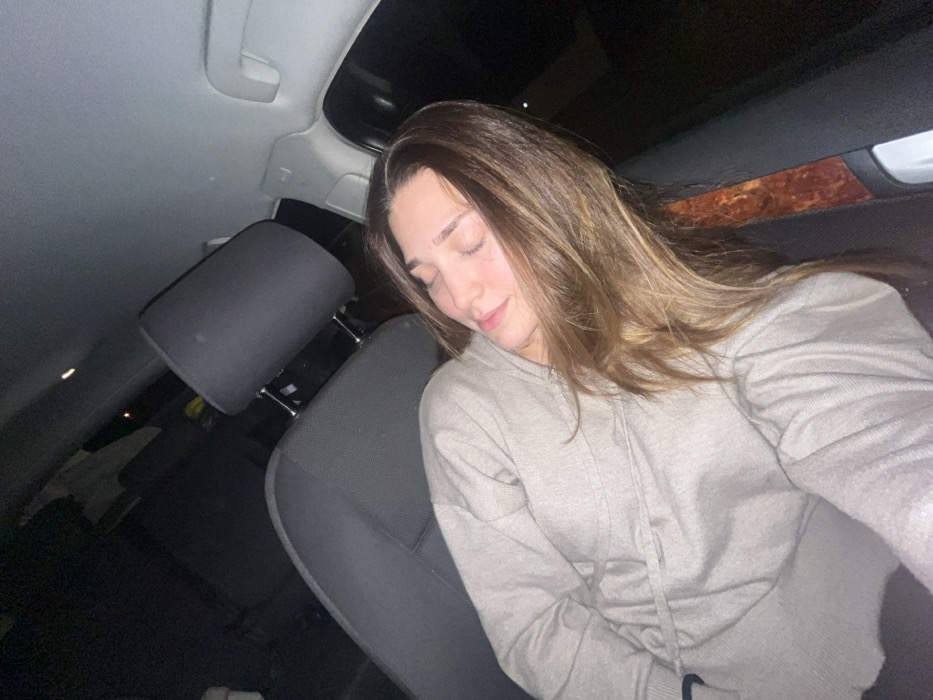 A young woman sleeps in a car seat, head resting against the headrest. She wears a light-colored hooded sweatshirt. Her long, light brown hair is parted to the side and falls over her shoulder. The car interior is dark, lit by dim ambient lighting. A sunroof is visible overhead. The seating material appears to be gray fabric.