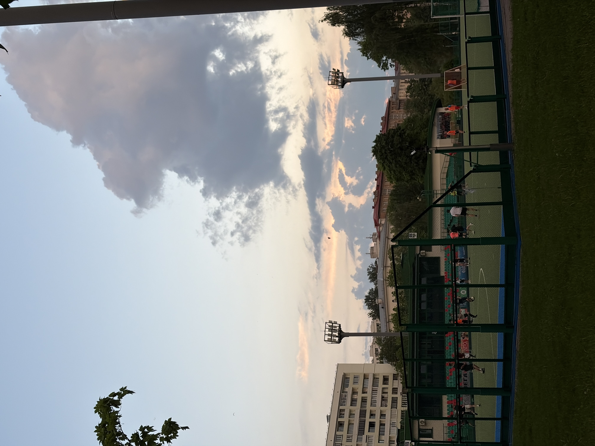 The image presents an outdoor scene in Minsk, Belarus, likely during the late afternoon or early evening. The sky is visible, featuring a mix of clear blue patches, wispy clouds tinged with hues of orange from the setting sun, and a large, dominant gray cloud formation.
Below the sky, the frame captures a recreational sports area. It includes fenced-in sports fields where people are engaged in some kind of ballgame, presumably soccer, or hockey. The players are wearing dark shorts and a variety of orange and black shirts. The sports facilities appear adjacent to an urban environment, with buildings partially visible, trees bordering the fields, and tall lampposts illuminating the activity areas. The background also shows apartment buildings with multiple stories. The grass surrounding the courts are very green.
Foreground elements include the edge of a building or structure in the top left corner, as well as the foliage of a tree branch.