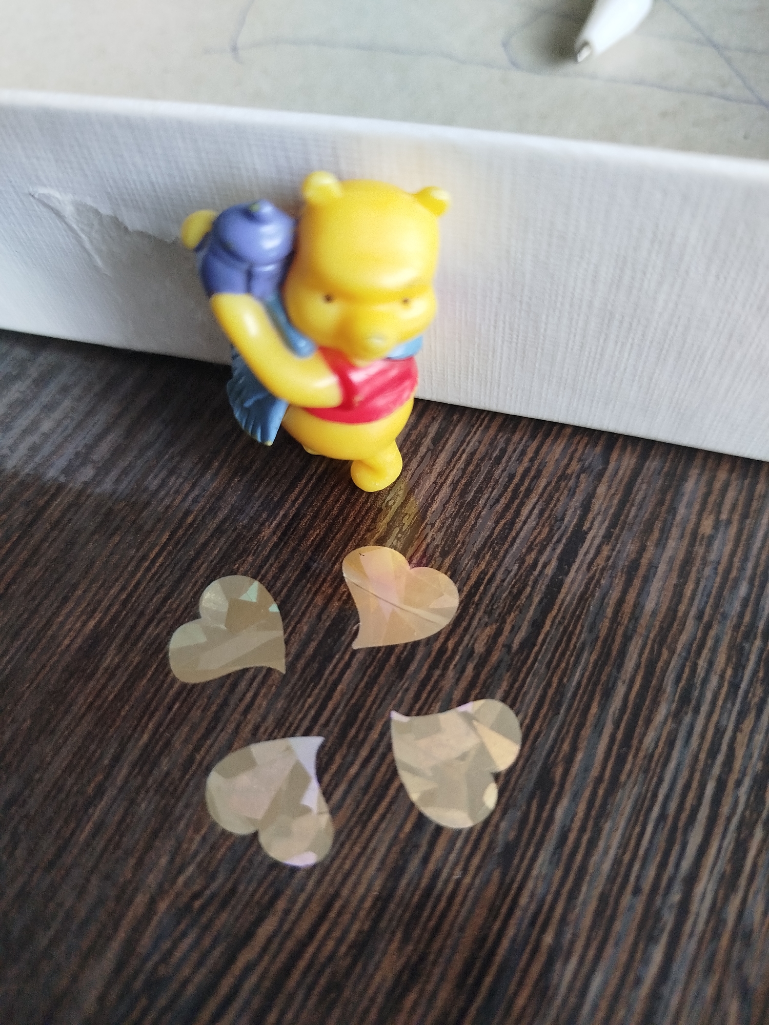 A small, yellow Winnie the Pooh figurine is positioned on a dark brown, wood-grain surface. The figurine is wearing a red shirt and blue pants, and it is holding a small blue object. In front of the figurine, there are four heart-shaped sequins scattered on the surface. The background features a white, textured surface. The image appears to be a close-up shot, with the focus on the figurine and the sequins. There is no identifiable location within the city of Крывасельскі сельскі Савет, Belarus.