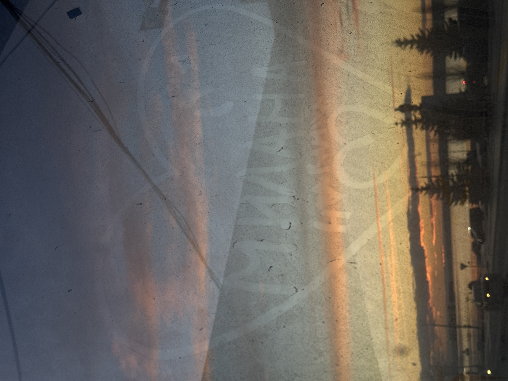The image, rotated 90 degrees clockwise, captures an outdoor scene viewed through a translucent and textured surface, possibly a dirty or frosted window. The time of day is either sunrise or sunset, indicated by the dramatic sky, which transitions from deep blue and grey on the left to vibrant orange, yellow, and pink tones on the right.
In the background, on what would be the horizon in a standard landscape orientation, dark silhouettes of conifer trees are visible, suggesting a forested edge or parkland. Behind these trees, faint outlines of buildings can be discerned, with a tiny red light glowing within one structure. Below the trees, along what appears to be a road or street, indistinct shapes resembling vehicles and glowing yellow lights (possibly streetlights or vehicle lights) are present.
The foreground and central area of the image are characterized by the window's surface, which exhibits numerous dust specks, smudges, and faint linear patterns, possibly scratches or reflections of power lines. Prominently displayed across the center is a translucent Cyrillic inscription within a larger, circular outline. The discernible text spells out "СНИМЕННОЕ" (SNIMENNOE), likely a watermark or a reflection of internal text, possibly meaning "photographed" or "shot." No specific human activity or identifiable individuals are depicted. The scene evokes a quiet, atmospheric moment in Votkinsk, Russia, during twilight hours.