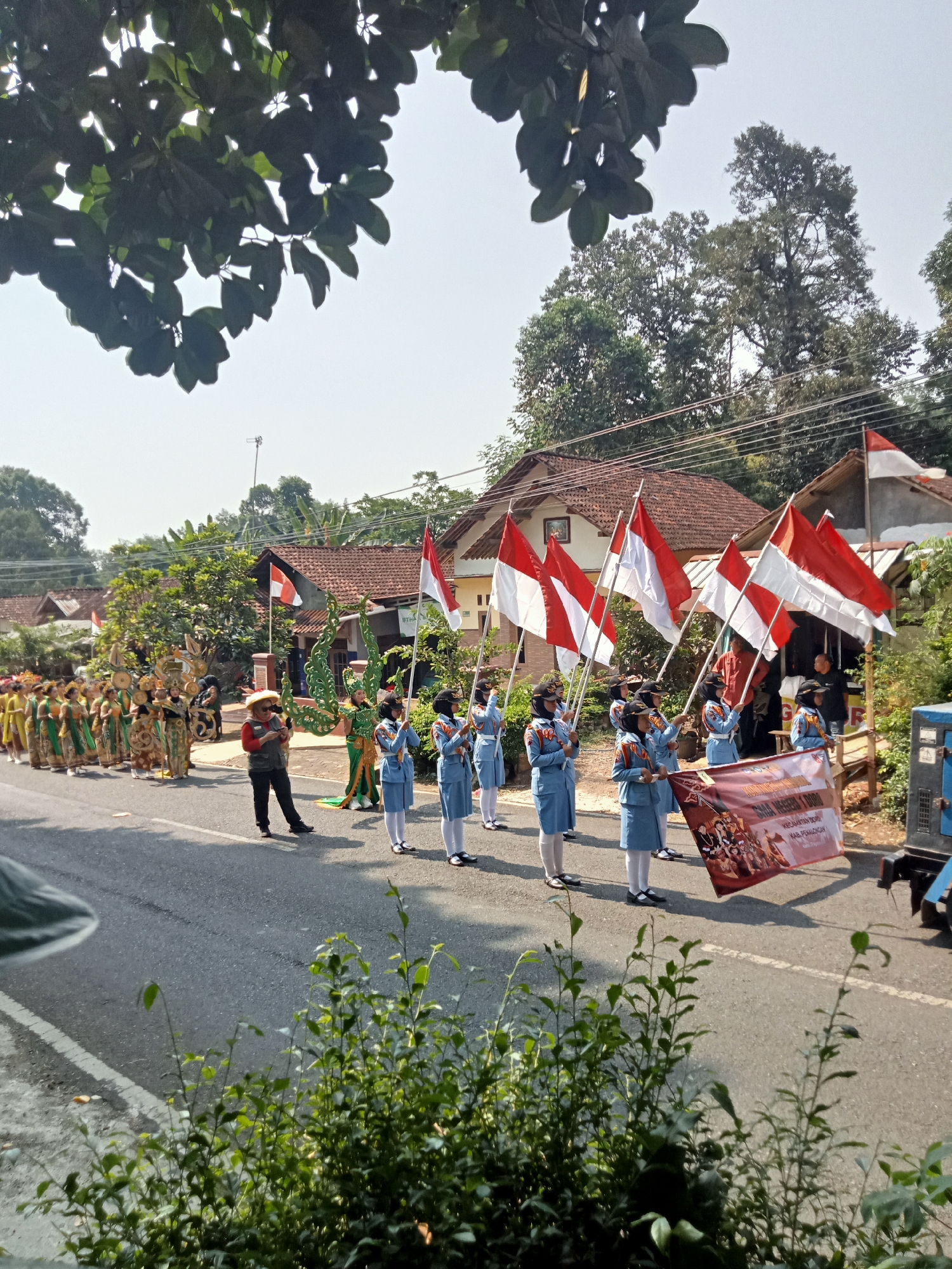 In a sunlit outdoor scene in Dororejo, Indonesia, a parade is in progress on a paved road. Several individuals, predominantly young women in light blue uniforms with black caps, are marching in formation. They are holding Indonesian flags, which are red and white, with the red stripe on top. Some of the flag bearers are also carrying a banner. Behind them, a group of people are dressed in elaborate, colorful traditional costumes, suggesting a cultural performance or procession. A person in a straw hat and red shirt appears to be directing or observing the parade. The backdrop consists of houses with red tile roofs and lush green trees under a clear sky. The road has a white line marking the edge. In the foreground, there are green leafy plants obscuring the bottom of the frame.