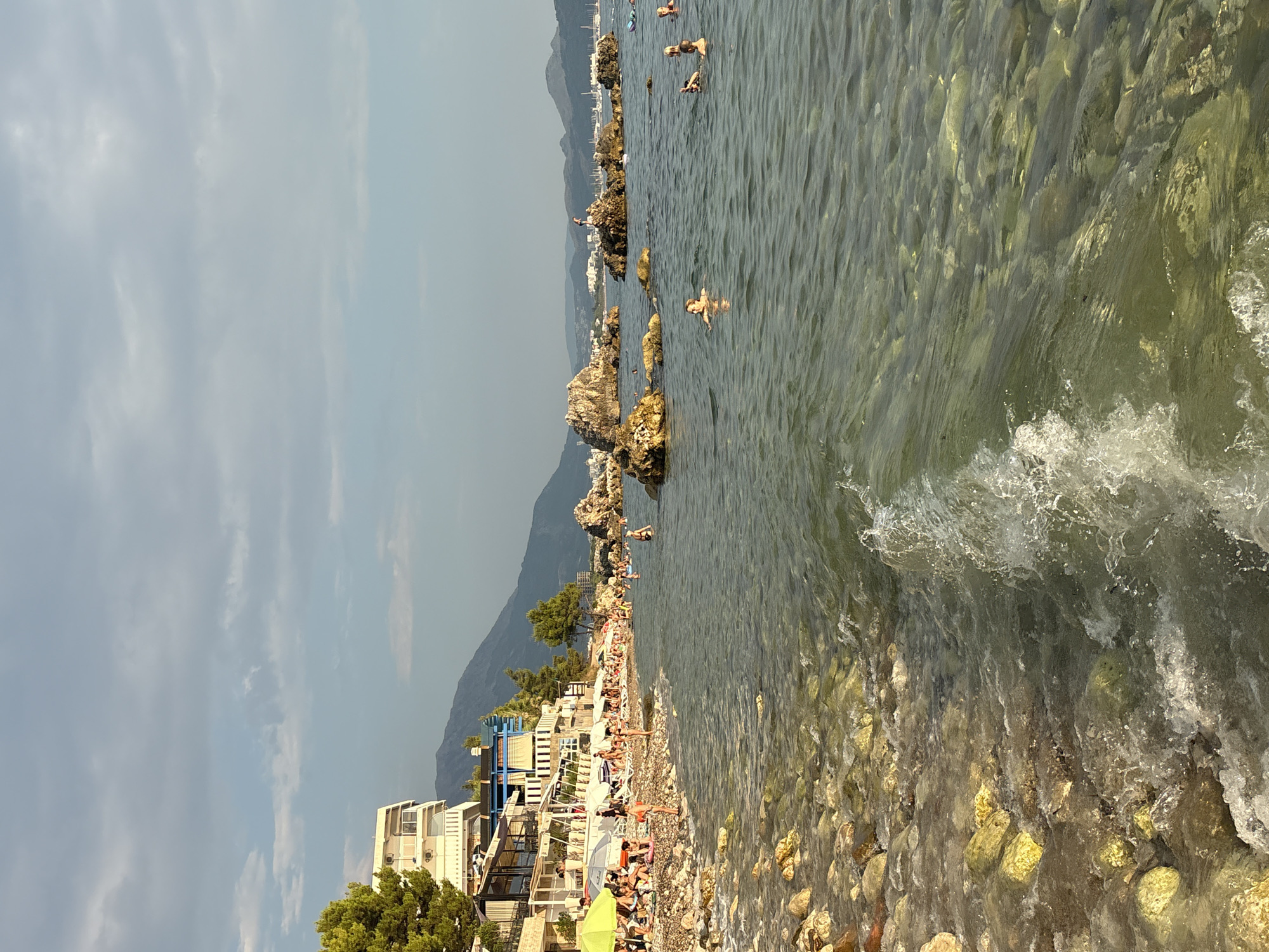 The image depicts a sunny day at a pebble beach in the city of Bar, Montenegro. In the foreground, clear, shallow water laps onto the shore, revealing submerged rocks and pebbles. A gentle wave breaks near the edge of the frame, creating white foam. The beach is lined with people relaxing on lounge chairs under umbrellas and towels. Several buildings, appearing to be hotels or apartments, are situated directly behind the beach, with awnings and balconies visible. Pine trees grow alongside the buildings. In the middle ground, several large rocks rise from the water, with a few people swimming and wading around them. The background features a hazy mountain range under a partly cloudy sky. The scene captures a typical summer day at a seaside resort.