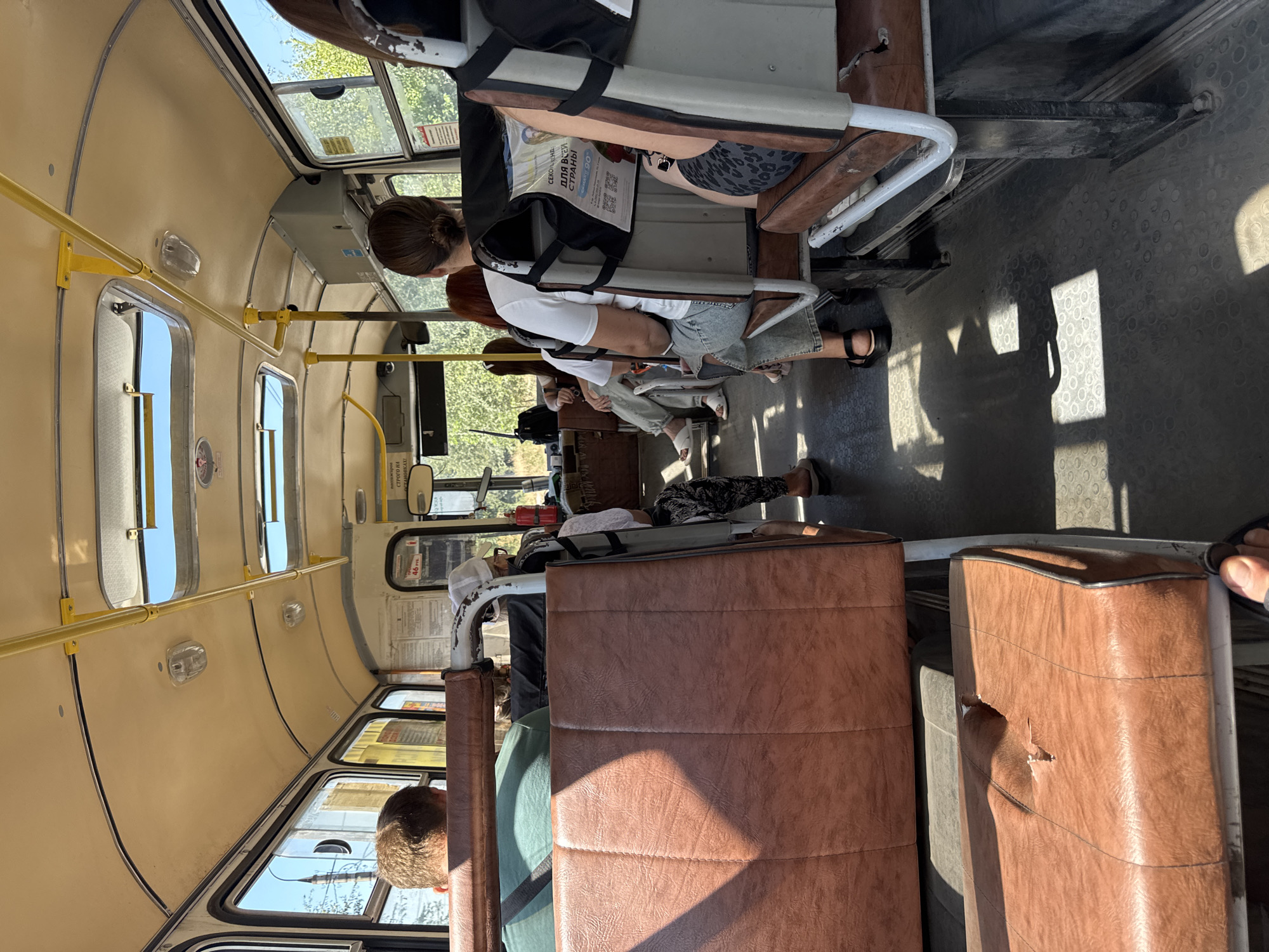 The image depicts the interior of a bus, likely in Shakhty City District, Russia, with several passengers seated. The bus has brown vinyl seating and a cream-colored ceiling with yellow handrails. Natural light streams in from windows and overhead skylights, casting shadows on the floor. Some passengers are visible from behind, engaged in travel. The overall impression is of a functional, somewhat aged public transport vehicle.