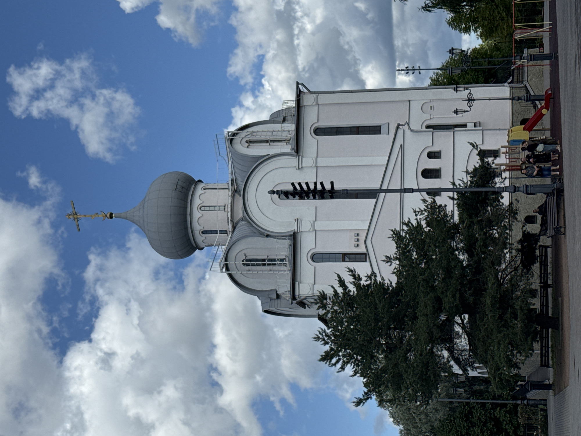 The image shows the Church of the Icon of the Mother of God of All Who Sorrow Joy, located in St. Petersburg, Russia. The church is a white building with a gray dome topped with a cross. A large evergreen tree is in the foreground to the left of the church. Several people are gathered near a playground with a red slide in front of the church. The sky is blue with white clouds.