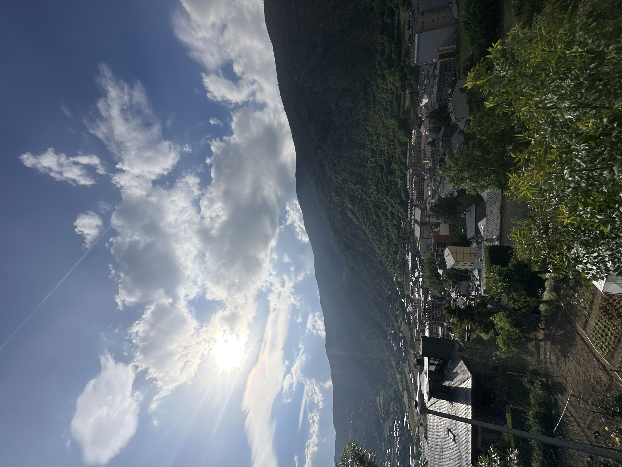 The image presents a scenic panoramic view of a mountainous landscape featuring a town, identified as Encamp, Andorra, under a bright, sunny sky. The photograph is taken during the daytime, likely late morning or early afternoon, as evidenced by the prominent sunburst and lens flare in the upper-mid portion of the sky.
The left half of the frame is dominated by a clear blue sky, dotted with numerous fluffy white clouds. A faint airplane contrail can be seen streaking across the upper left quadrant. The sun's intense rays create a starburst effect, illuminating the clouds and adding a vibrant warmth to the scene.
On the right side, a massive, steep mountain rises majestically, densely covered from base to peak with lush green forest. Its verdant slopes provide a dramatic backdrop to the settlement below. Nestled at the foot of this mountain and extending into the mid-ground is the town of Encamp. Its buildings feature a mix of architectural styles, with many roofs appearing dark, possibly made of slate or tiles, characteristic of mountainous regions.
In the foreground, towards the lower-left, are trees and bushes with rich green foliage, partially framing the view. Below these trees, a cultivated garden or field with discernible rows of plants suggests local agricultural activity. The overall impression is one of natural beauty and tranquility, showcasing a community integrated into a grand mountainous environment. There are no visible people or discernible text within the image.