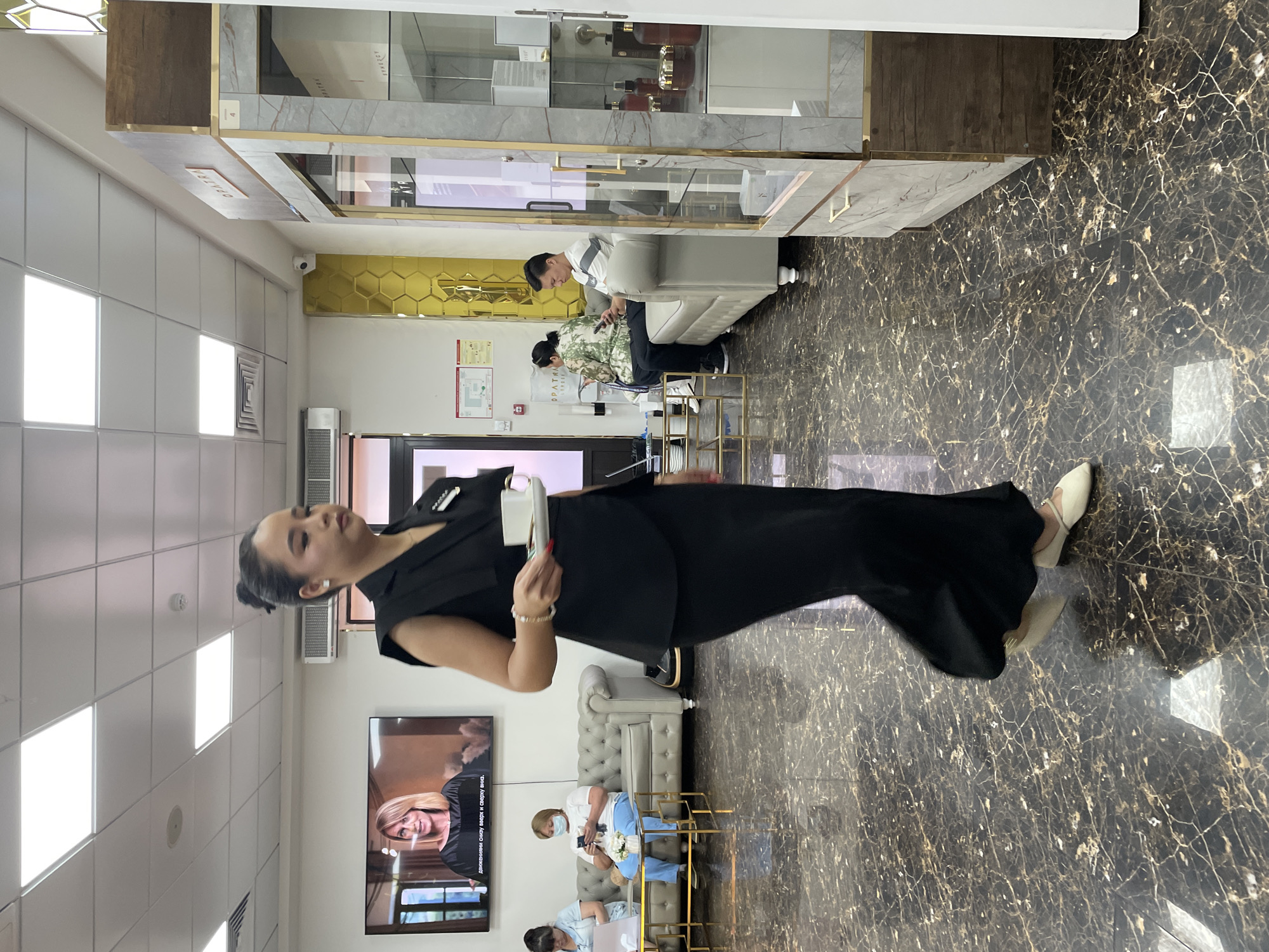 The image depicts the interior of a modern establishment, likely a beauty salon or a spa, located in Almaty, Kazakhstan. The foreground is dominated by a woman walking from left to right, dressed in a black sleeveless vest over a black skirt and cream-colored flat shoes. She is carrying a small white cup and saucer in her right hand, suggesting she is serving a beverage. Her hair is pulled back in a neat bun, and she wears subtle jewelry.
In the background, several people are seated on a light grey sofa. On the left, a woman in a light blue top and dark pants is looking at her phone. To her right, a woman in a white t-shirt and blue pants is also looking at her phone, with a white face mask partially covering her lower face. Further to the right, a man in a striped t-shirt is seated on the sofa, looking down at his phone.
The environment features a polished dark marble floor with a light-colored veining pattern. The walls are painted white, with a large flat-screen television mounted on the left wall displaying a woman's face. To the right, a tall display cabinet with glass doors showcases various cosmetic products. A golden hexagonal pattern adorns a section of the wall behind the sofa, adding a touch of opulence to the space. Fluorescent ceiling lights illuminate the room. The overall impression is one of a clean, well-lit, and contemporary commercial space.