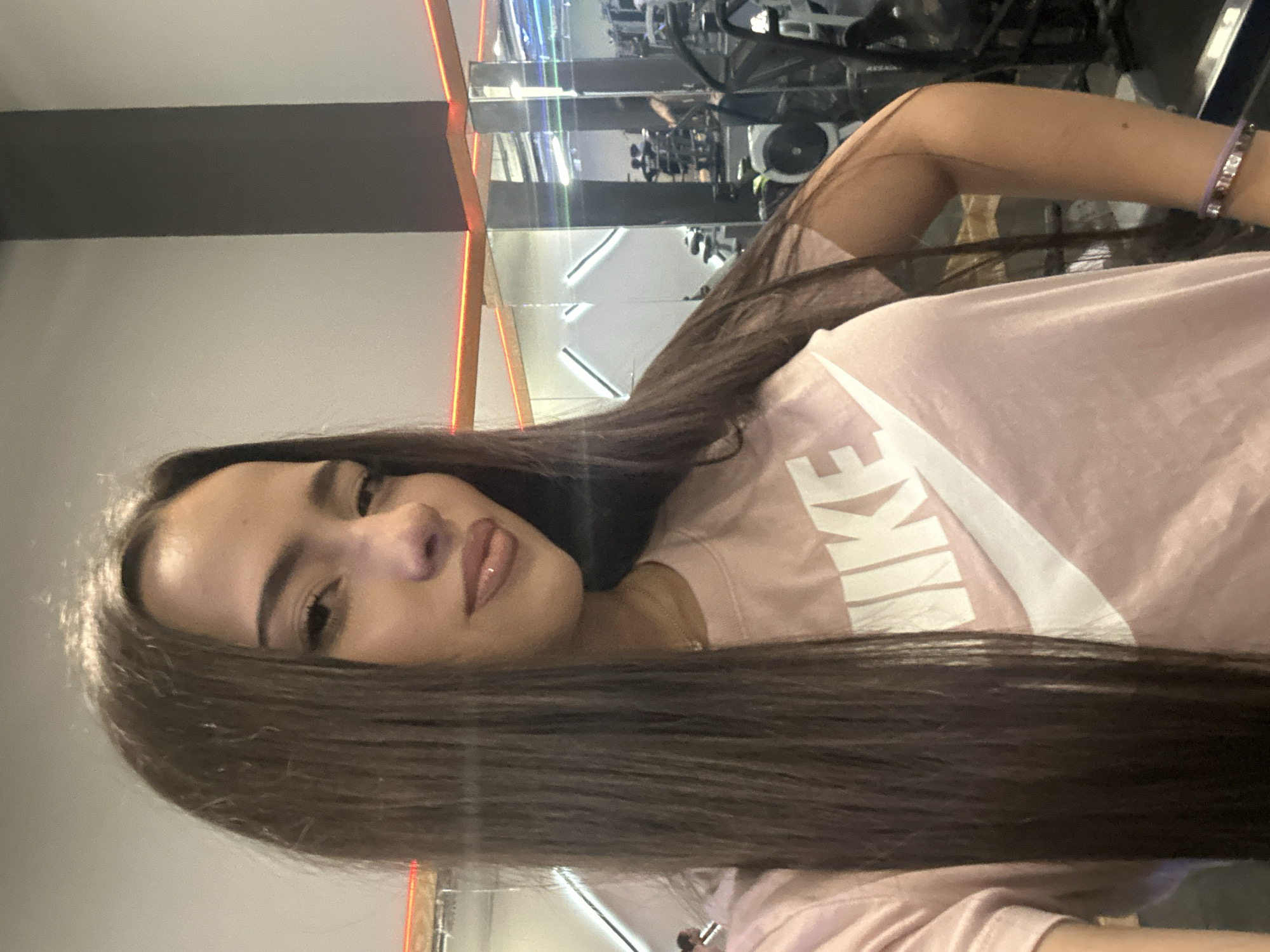 A selfie captures a woman with long, dark hair wearing a pink Nike t-shirt. She's in a gym, as indicated by the exercise equipment visible in the mirrored background. The location is the city of Unknown, Unknown.