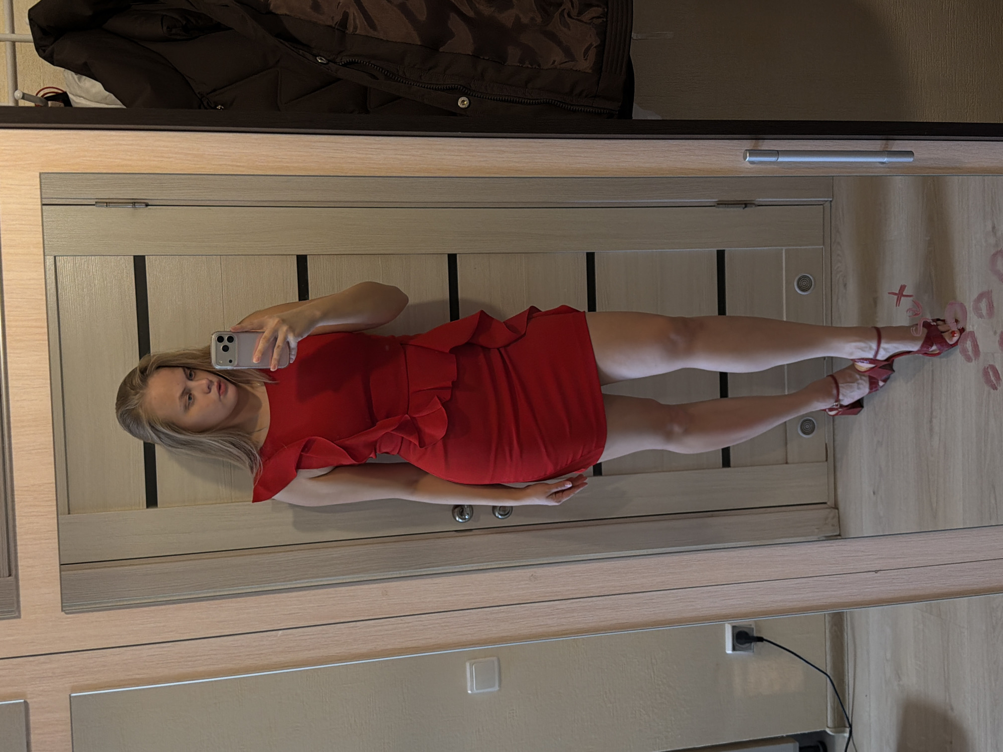 A young woman is reflected in a full-length mirror. She is wearing a red dress and red high-heeled sandals. She is holding a cell phone in her hand, presumably taking a selfie. The reflection also shows a closet with a jacket hanging inside, a light switch, and an electrical outlet. There are lipstick marks on the wall next to the woman's feet. The location is Izhevsk, Russia.