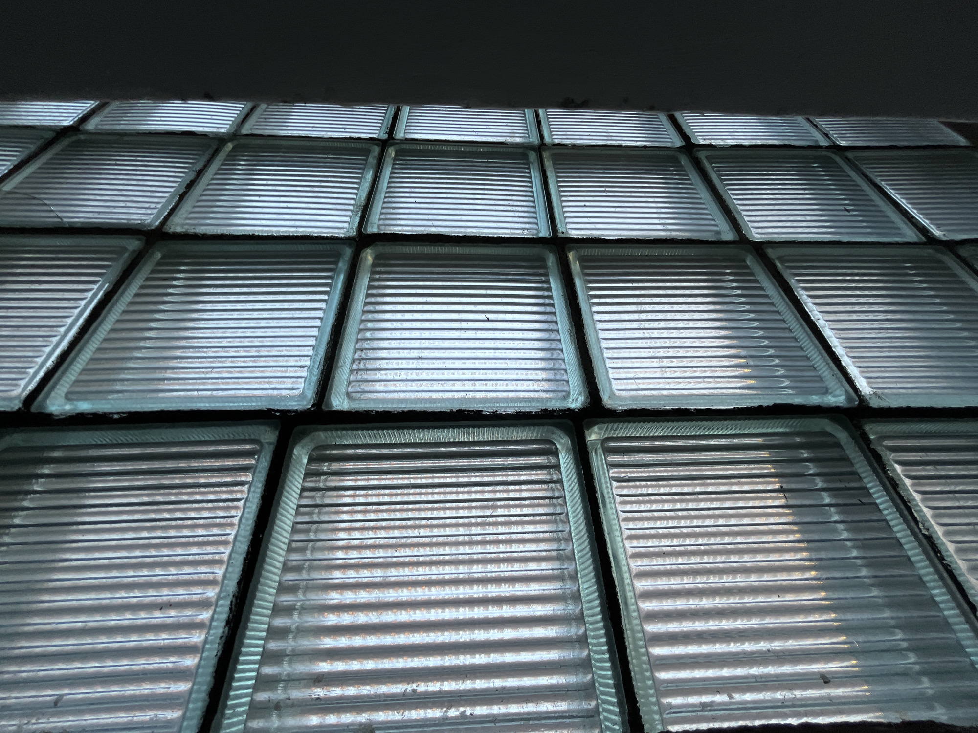 The image depicts a close-up, low-angle view of a wall constructed from numerous rectangular glass blocks. These blocks are translucent and feature a ribbed, horizontally textured surface, creating a pattern of parallel ridges and troughs. They are arranged in a grid formation, appearing in multiple rows and columns, with dark grout or sealant filling the spaces between them.
The lighting strongly emphasizes the texture of the glass blocks. Light appears to be filtering through or reflecting off their surfaces, creating distinct horizontal bands of highlights and shadows on each block. This play of light gives the blocks a shimmering, almost metallic appearance in some areas, while others remain more subdued. The top portion of the image is dominated by a dark, indistinct area, possibly a ceiling or an overhead structure, which contrasts with the illuminated glass blocks below.
No specific activity or event is taking place; the image is a static architectural detail. The lighting suggests an indoor setting or a sheltered outdoor area, likely during the daytime, with ambient light illuminating the structure. There are no discernible location-specific cues beyond the typical appearance of a glass block wall, commonly found in various building types for light and privacy. No emotions are explicitly conveyed, and there is no visible text in the image.