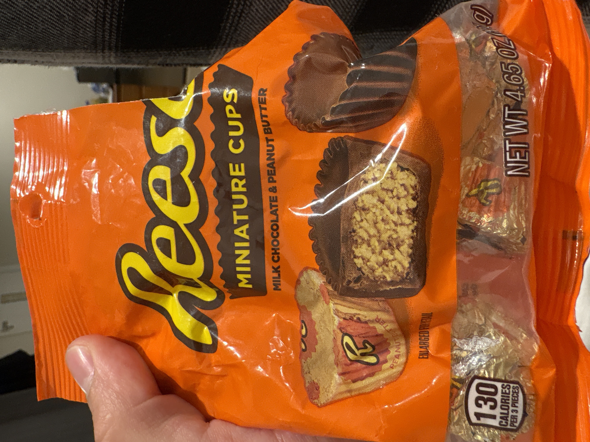 The image shows a hand holding a crinkled, orange-colored bag of Reese's Miniature Cups. The bag is made of a shiny plastic material, and its top left corner appears slightly torn or open, suggesting it has been handled or recently opened.
The dominant feature on the bag is the large, yellow, and brown "Reese's" logo. Below it, the text "MINIATURE CUPS" is visible, along with a description "MILK CHOCOLATE & PEANUT BUTTER". The packaging prominently displays illustrations of the candy: one whole Reese's cup, another cut in half to show the peanut butter filling, and a third unwrapped from its paper liner, revealing the rippled chocolate cup. Text below these illustrations reads "ENLARGED FOR DETAIL".
On the right side of the bag, a clear plastic strip reveals several individually foil-wrapped Reese's miniature cups inside. Vertical text on this strip indicates "NET WT 4.65 OZ (131g)". Further down, a white oval graphic with "130 CALORIES PER 3 PIECES" is clearly visible, providing nutritional information.
The hand holding the bag is in the lower-left portion of the frame, with the thumb partially obscuring some of the orange plastic. The background is dark and somewhat blurry, suggesting an indoor setting, possibly a piece of furniture like a couch or chair, given the textured fabric visible at the top. There are no other people visible, and no specific time of day or weather can be discerned from the image.