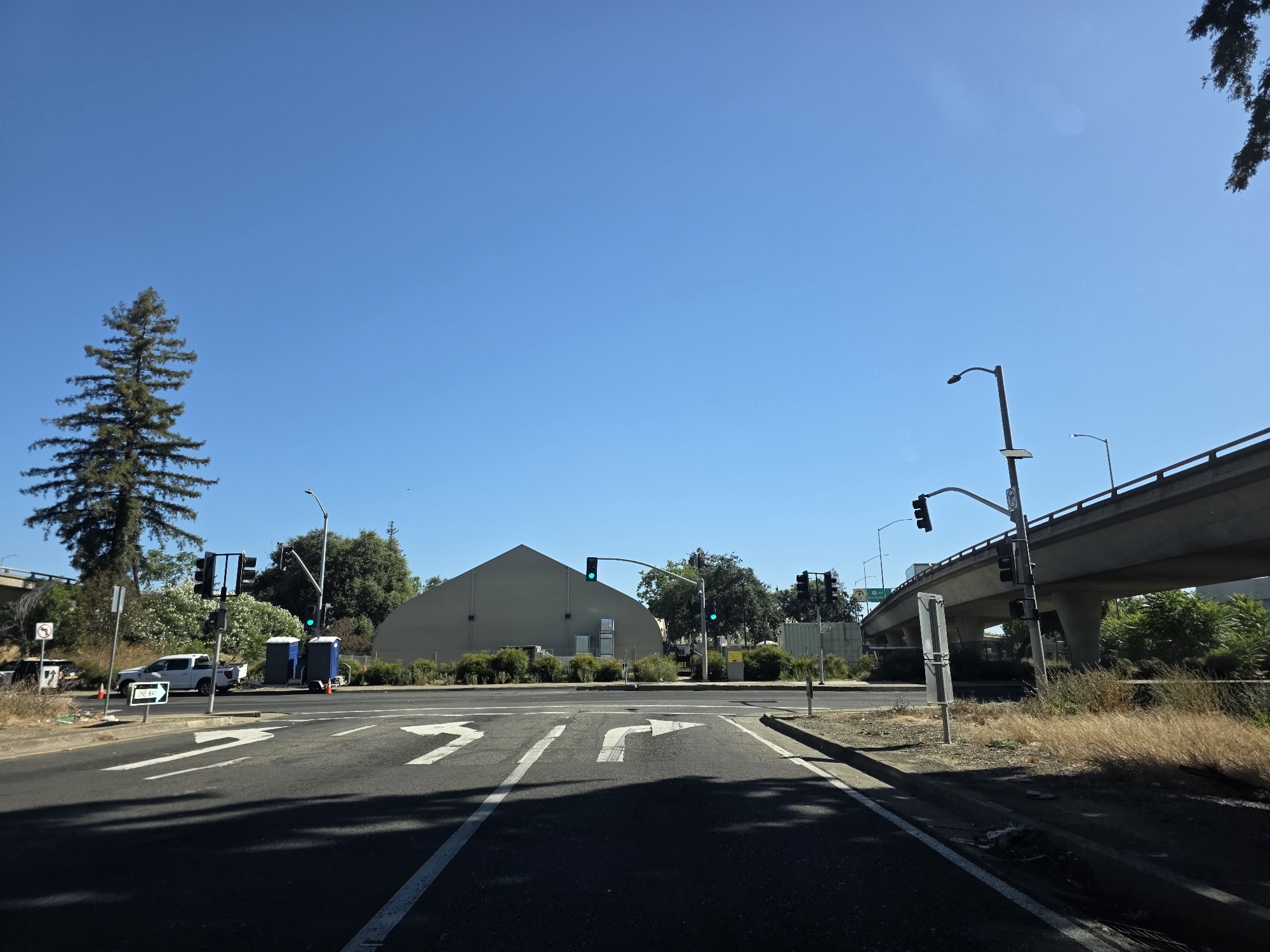 This media file depicts an outdoor scene at a street intersection under a clear blue sky, suggesting a sunny day. The view is from the perspective of someone on the road, looking forward. In the foreground, the asphalt road is marked with white directional arrows indicating lanes for going straight, turning left, and turning right.
The mid-ground features an intersection with traffic lights, some of which are illuminated green. Several portable restrooms are visible, placed on a paved area near the roadside. A white pickup truck is parked nearby. To the left, a tall evergreen tree stands prominently against the sky. Behind the portable restrooms and truck, a large, light-colored building with a distinctive triangular roof is visible. Further back, there is dense foliage and more trees.
In the background, a concrete overpass or highway bridge looms overhead, with cars visible on it. The setting appears to be urban or suburban, possibly in Sacramento, California, given the provided location context. The dry, golden grass along the side of the road hints at a warm climate. There is no indication of any specific activity or event taking place, beyond the normal flow of traffic at an intersection. The overall mood is neutral, reflecting a typical daytime scene. There is some text visible on a sign that says "ONE WAY" with an arrow pointing to the right. Another sign, partially obscured, appears to be a traffic regulation sign.