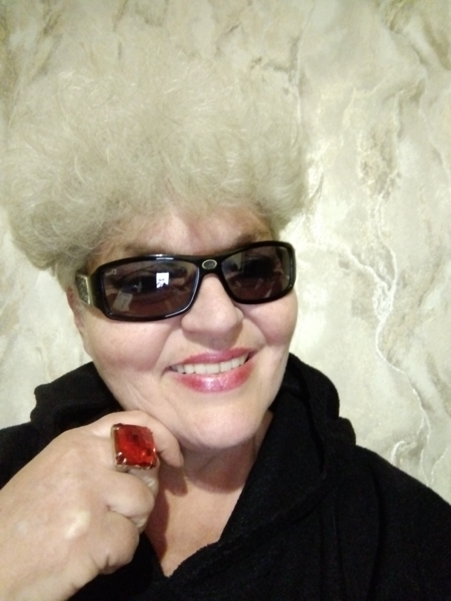 An older woman with white, fluffy hair poses for a selfie in an indoor setting. She wears large, dark-framed sunglasses and a bright red, square ring on her left hand. She is smiling, showing her teeth, and has lipstick on. She is wearing a dark-colored hooded garment. The background is a wall with a light, marble-like pattern. The location is assumed to be somewhere in Horishni Plavni, Ukraine.