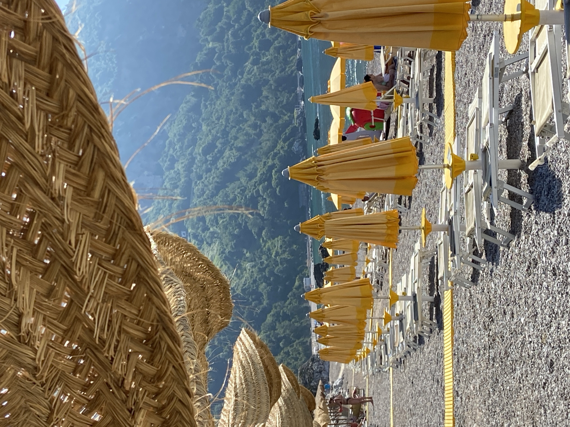 This is a low-angle, vertical, eye-level shot of a beach in Prijevor, Montenegro, on a sunny day. The foreground is dominated by rows of white lounge chairs and closed, yellow beach umbrellas on a pebbly beach. The umbrellas are made of fabric stretched over a metal frame, with a yellow border. In the midground, a few people are visible on the beach. To the left, a person in a black bikini stands facing away from the camera. Further back, another person sits on a lounge chair. In the background, the calm, blue sea meets a lush, green mountainside, dotted with buildings and rocks. Above the beach umbrellas, thatched-roof beach umbrellas hang, providing shade. The scene suggests a typical beach resort environment with amenities for visitors.