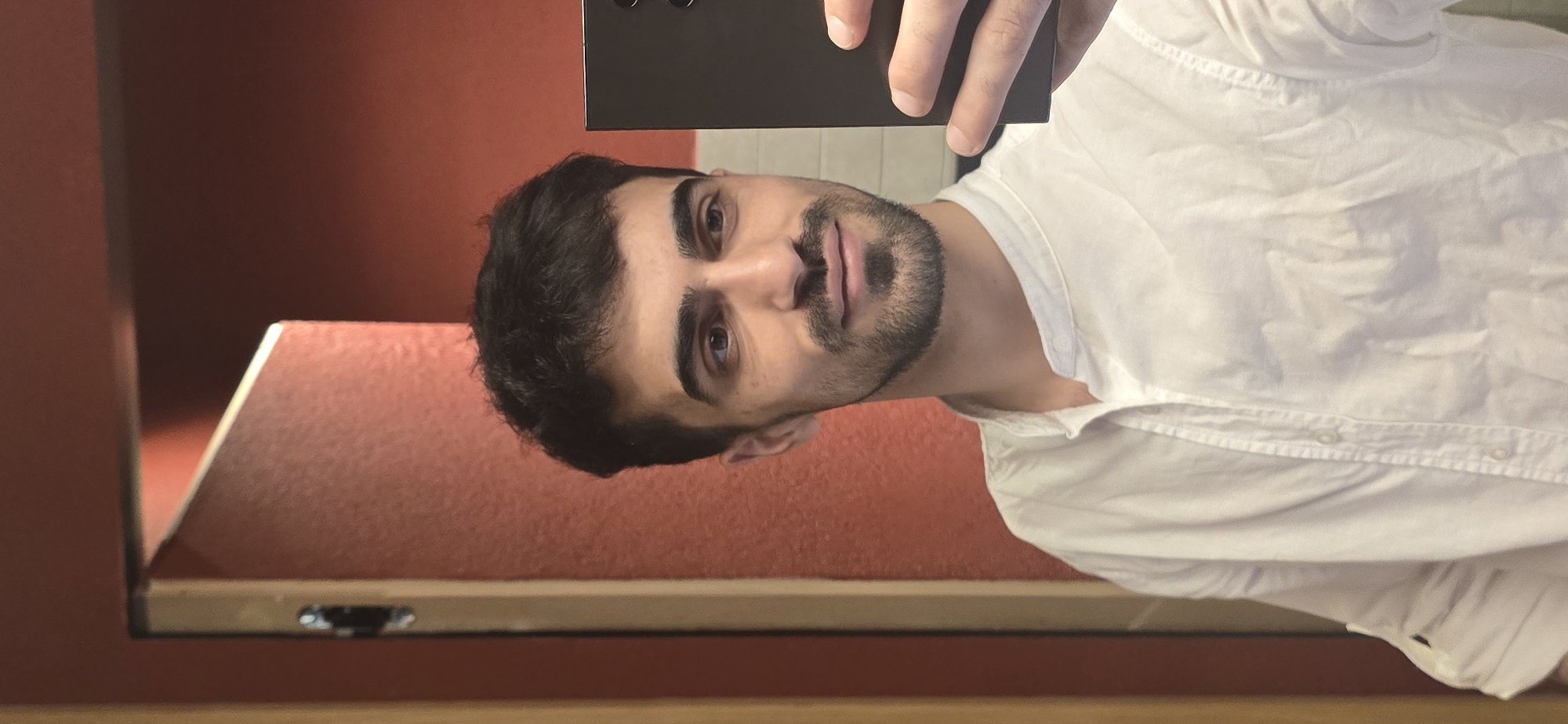A man is taking a selfie in what appears to be a room with dark red walls. He is holding a black smartphone or similar device. The man has dark hair, brown eyes, and a beard. He is wearing a white button-down shirt. The bottom of the image shows what looks like the edge of a table or shelf, with a possible power outlet. There is no readily apparent evidence of Yerevan, Armenia.