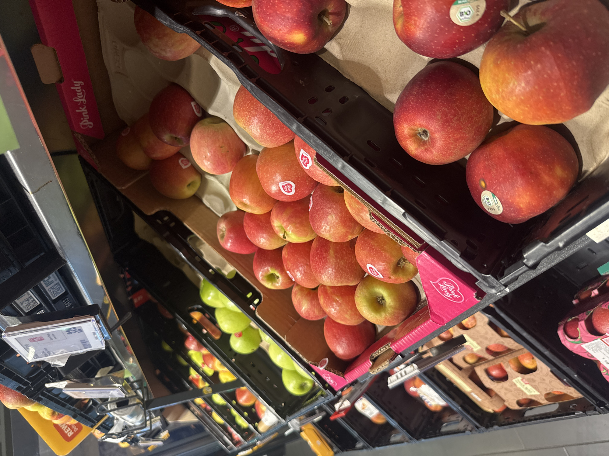 A close-up, angled shot shows a display of apples in a grocery store. The apples are a mix of red and green varieties, with some showing pink and yellow hues. They are arranged in black plastic crates, some of which are lined with brown paper. One crate prominently displays a pink insert with the words "Pink Lady" in white lettering. Several apples have round, white stickers with green accents, one of which reads "BIO." The lighting is bright, suggesting an indoor retail environment. In the background, shelves are visible with more produce and price tags. The overall impression is of a fresh produce section in a supermarket.