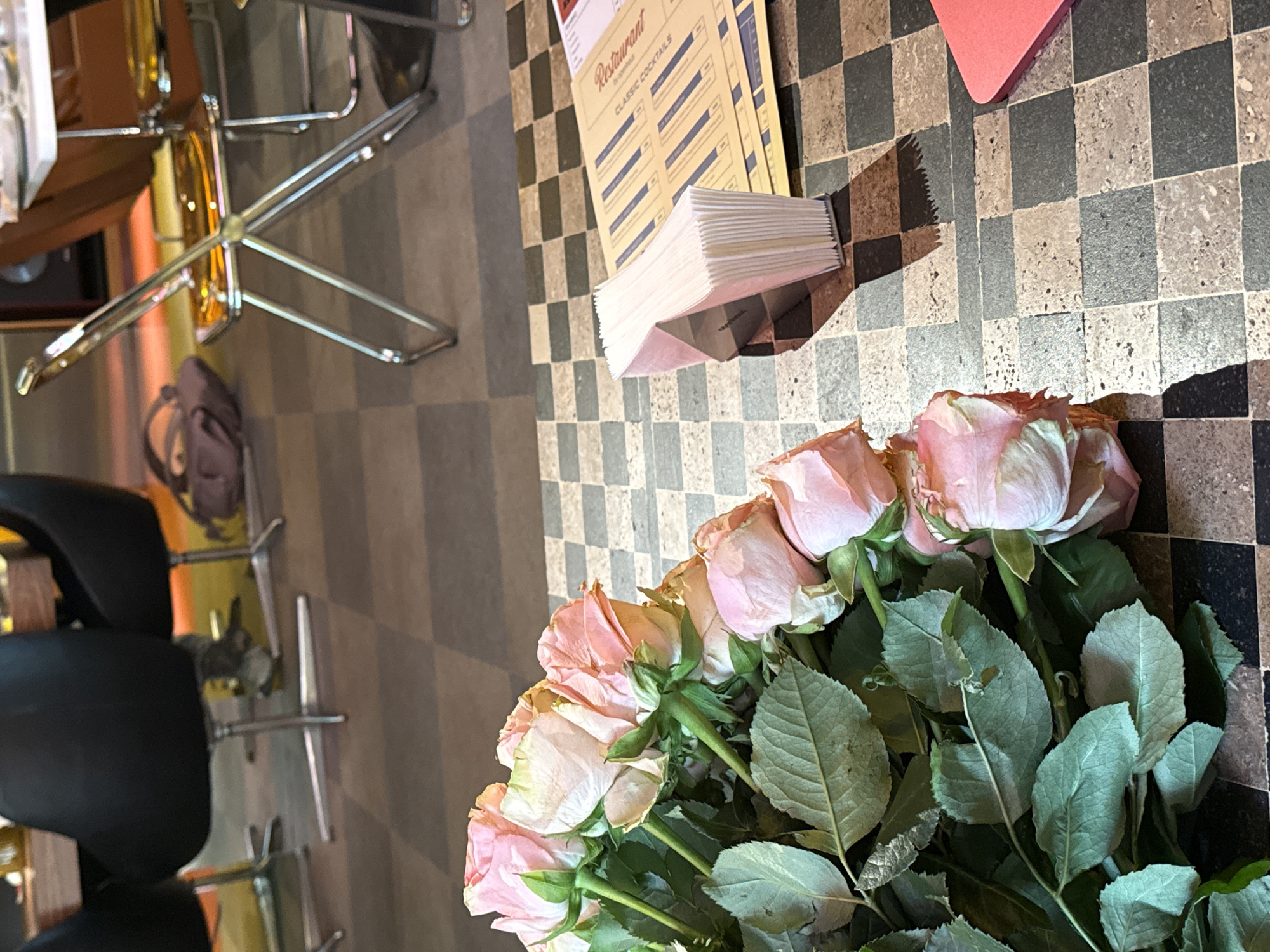 The image shows a restaurant table in a Yerevan, Armenia establishment. The table is covered with a checkered pattern. Arranged on the table are a bouquet of pink roses, restaurant menus, a stack of napkins, and a red heart-shaped object. A chair is visible near the table.