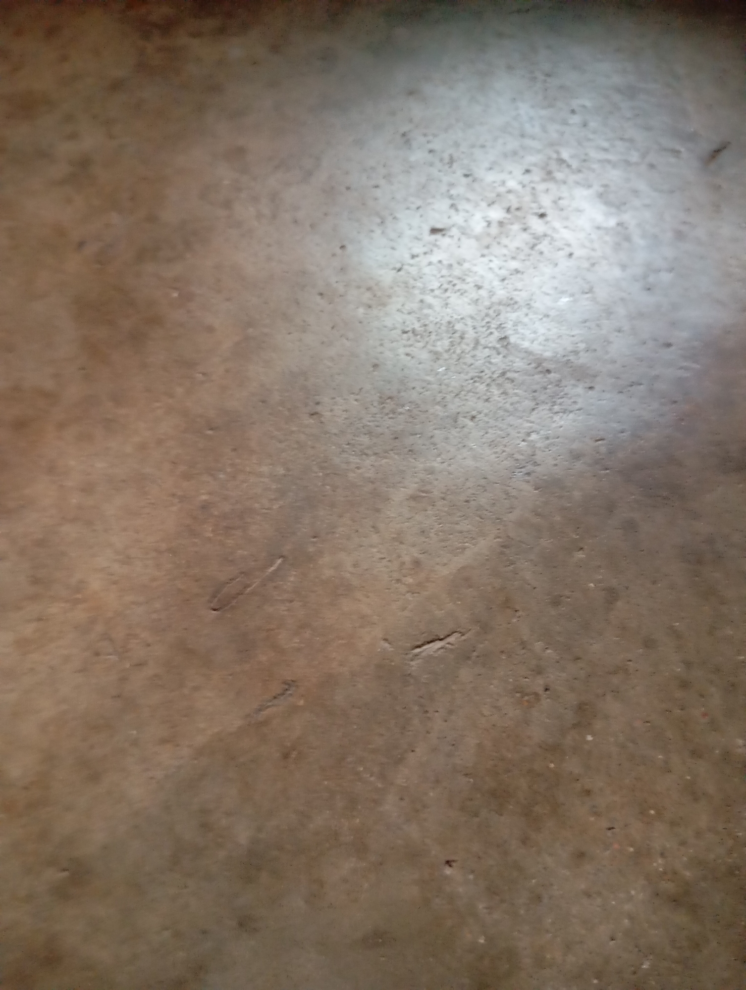 The image displays a close-up, overhead view of a textured, brown and gray concrete surface. The surface appears to be a floor, possibly outdoors or in an unfinished interior space. There are several subtle indentations and scratches on the concrete, with a brighter, reflective area on the upper right quadrant, suggesting a light source. The texture is rough and uneven, with small, dark specks scattered throughout. There are no people or discernible objects in the frame. The scene is devoid of any specific identifying markers that would allow for precise localization within Jalingo, Nigeria.