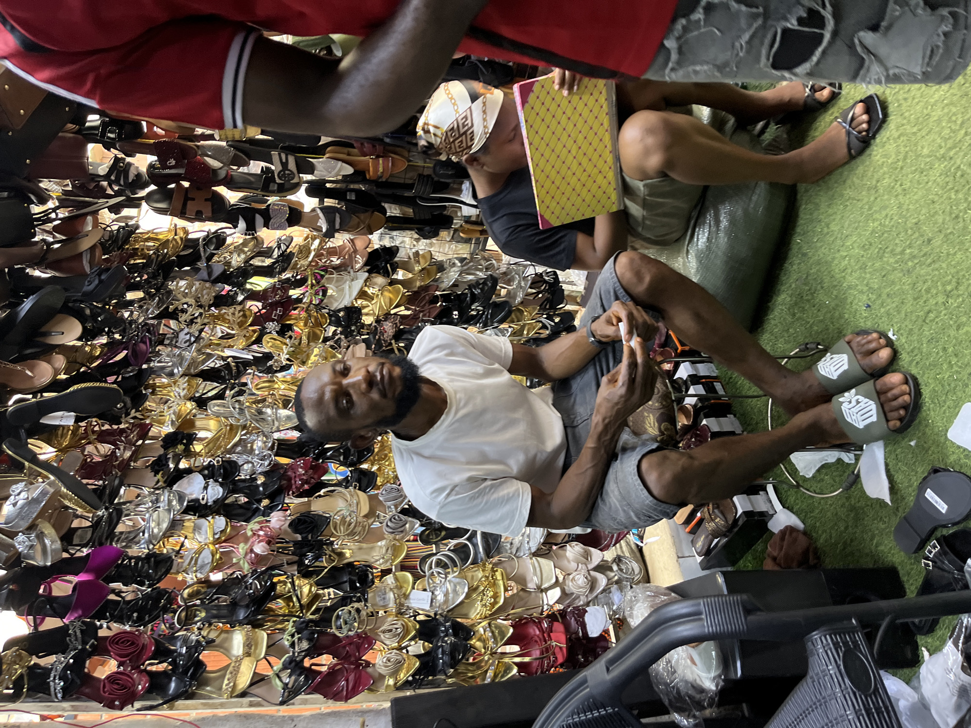 The image shows a street-level shoe stall in Abuja, Nigeria. The stall is packed with numerous pairs of women's sandals and heels, displayed on shelves and in piles. Colors range from vibrant reds and purples to metallics like gold and silver, alongside classic black and neutral tones. Many of the shoes feature decorative elements like bows, flowers, and intricate straps.
Three people are visible in the scene, all appearing to be engaged with the merchandise or the business of the stall. In the foreground, a man with a beard, wearing a white t-shirt and grey shorts, is seated and looking towards the camera with a neutral expression. His right hand is resting on a pile of shoes. To his right, another person, likely a man, is also seated and wearing grey shorts and a black t-shirt. He is holding a yellow and pink patterned book or folder. Another person, wearing a red shirt with white and black trim, is partially visible at the top of the frame, their arm extended.
The setting is a bustling market environment, suggested by the dense display of goods and the presence of multiple individuals. The ground appears to be covered in artificial green turf. The lighting suggests it could be daytime. The overall impression is one of a busy commercial space focused on retail sales.
