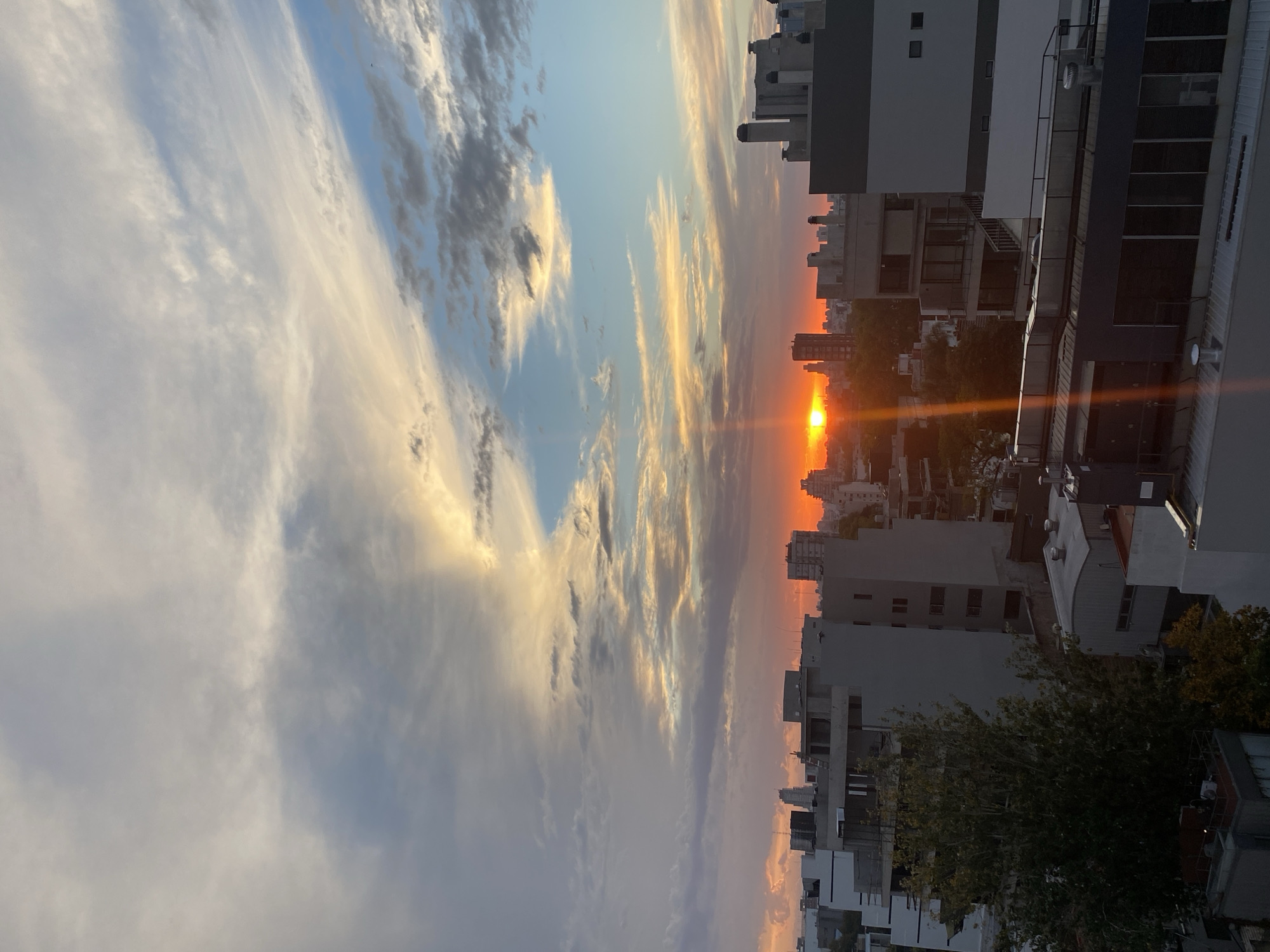 The image depicts a sunset in a city, likely Buenos Aires, Argentina, as indicated by the architectural style of the buildings. The sun is positioned low on the horizon, casting an orange and red glow over the cityscape. Buildings of varying heights are silhouetted against the bright sky. The sky itself is a mix of blue and orange hues, with clouds scattered throughout. The foreground includes rooftops and tree foliage. The perspective suggests the image was taken from an elevated position, such as a balcony or rooftop. There are no people visible in the image.