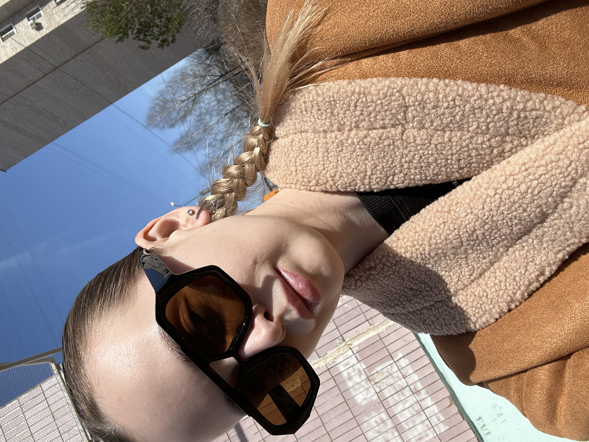 A medium shot photograph shows a woman with a blonde braid wearing sunglasses and a brown jacket with a beige, sherpa-lined collar. The shot is taken outdoors, with a building and trees visible in the background and a blue sky. The woman is wearing a black top. The picture was taken in the city of Chaikovsky, Russia.